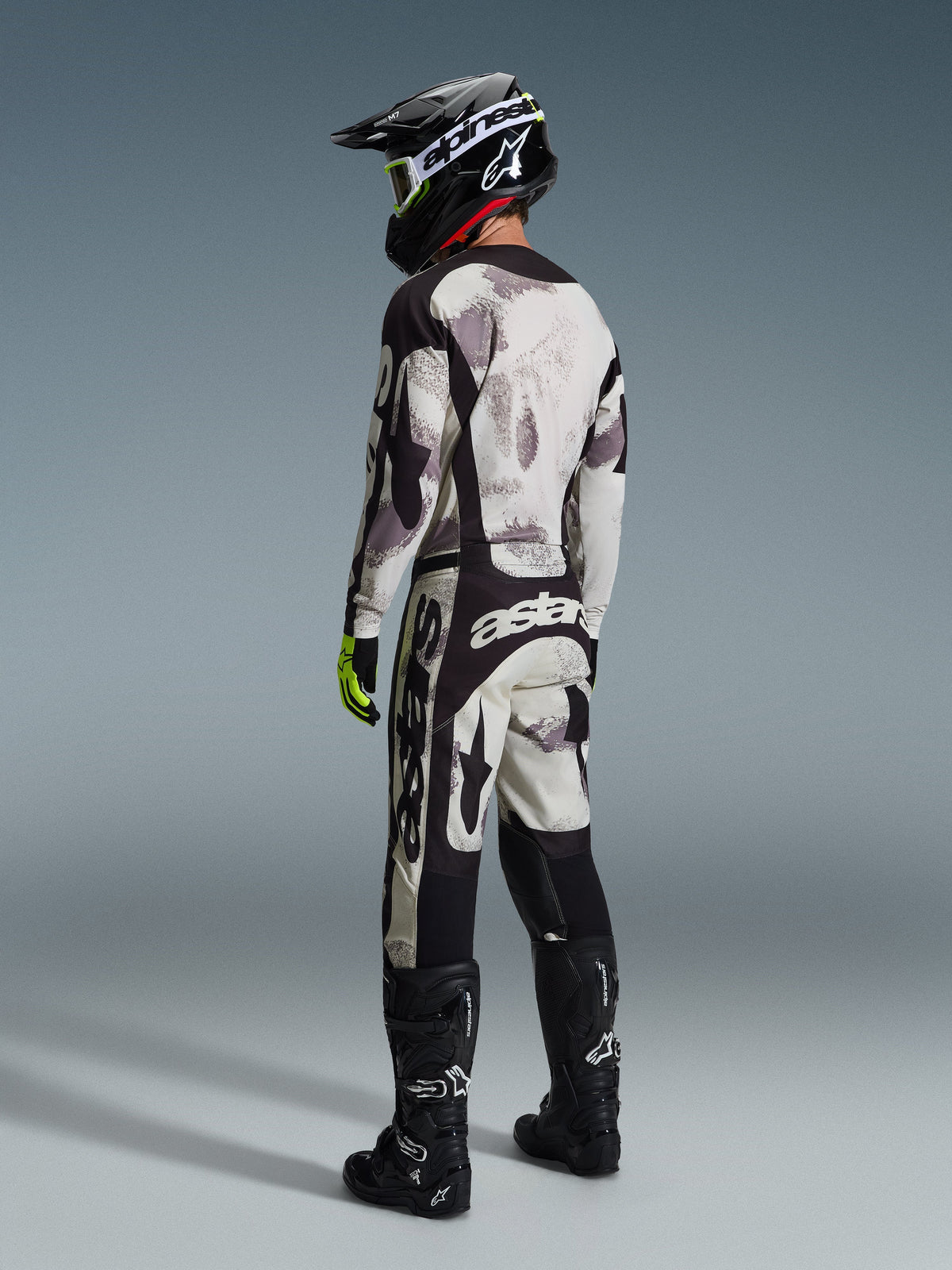 Motocross-Fahrer in kompletter Rennausrüstung, 2026 Racer Lahnd Trikot und passende Hose in Sand Camo, mit grau-schwarzem Camouflage-Muster und markanten Alpinestars-Logos, schwarzer Helm, schwarze Stiefel und neongelbe Handschuhe, Rückansicht.