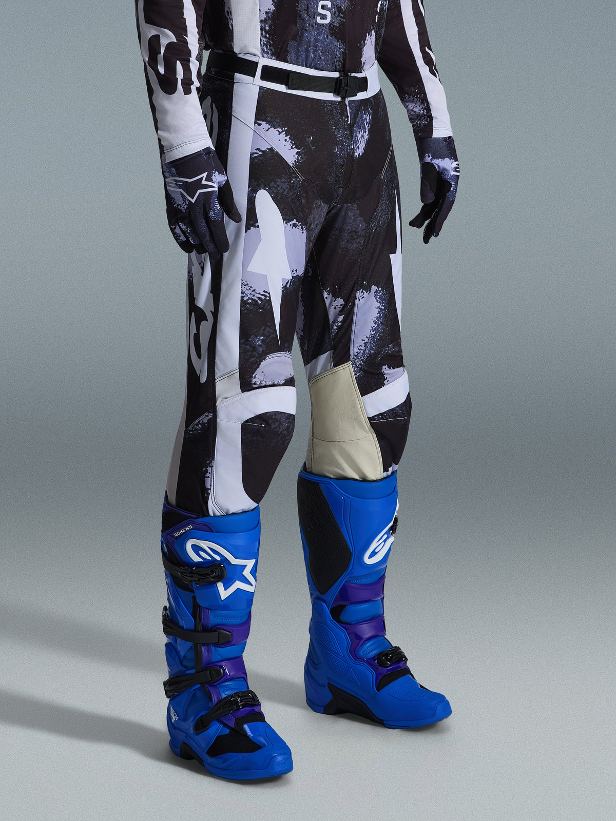 Alpinestars 2026 Racer Lahnd Hose, Motocross-Hose, grau-schwarzes Iron Camo Muster, getragen mit leuchtend blauen Rennstiefeln und passenden Handschuhen, mit weißen Einsätzen und Knie-Innenseiten aus Ziegenleder, leistungsorientierte Offroad-Ausrüstung