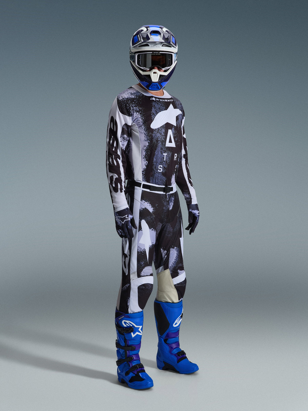 Motocross-Fahrer in voller Ausrüstung mit Alpinestars 2026 Racer Lahnd Hose, Iron Camo und grauem Muster, passendes Trikot, leuchtend blaue und lila Motocross-Stiefel, blau-weißer Helm, professionelles Offroad-Rennoutfit