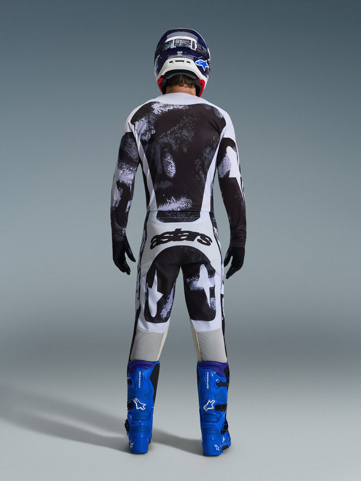Alpinestars 2026 Racer Lahnd Hose, Motocross-Hose, grau-schwarzes Iron Camo Muster, Rückansicht eines Fahrers in voller Rennausrüstung mit passendem Trikot und blauen Stiefeln, entwickelt für Offroad-Performance.