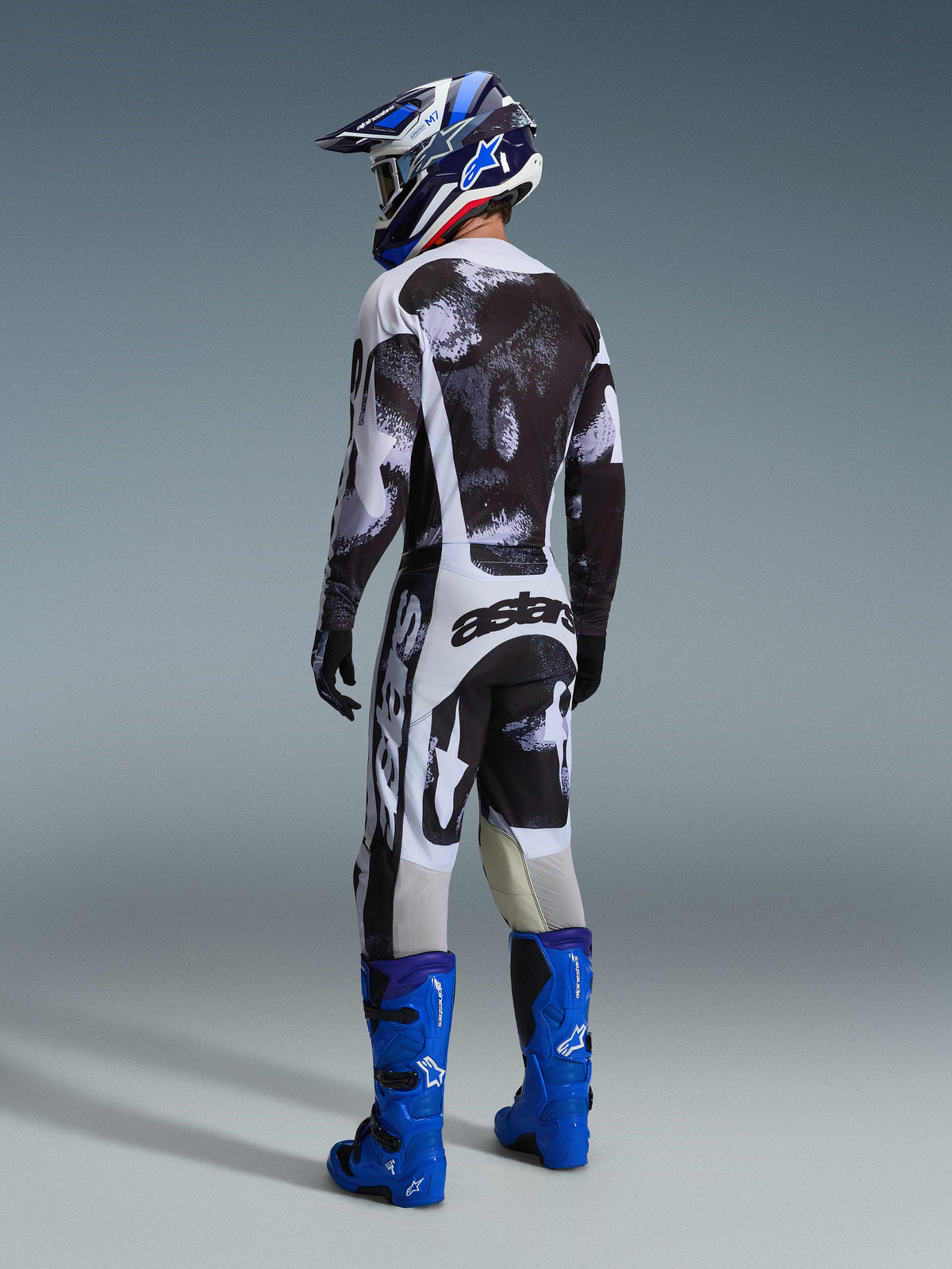Motocross-Fahrer in kompletter Rennausrüstung von hinten, 2026 Racer Lahnd Trikot und Hose in Iron Camo, schwarz-grau-weißes abstraktes Muster, leuchtend blaue Alpinestars Stiefel, blau-weißer Helm, abgestimmtes Outfit für den Offroad-Rennsport