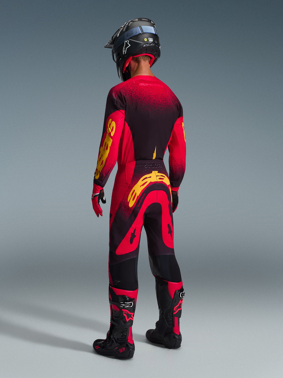 Motocross-Fahrer in der Rückansicht mit Alpinestars 2026 Supertech Scenz Hose in Rot-Schwarz-Gelb, passendes Jersey mit auffälligem gelbem Branding, schwarz-rote Motocross-Stiefel, schwarzer Helm, komplettes Profi-Rennausrüstungsset für Offroad-Performance.