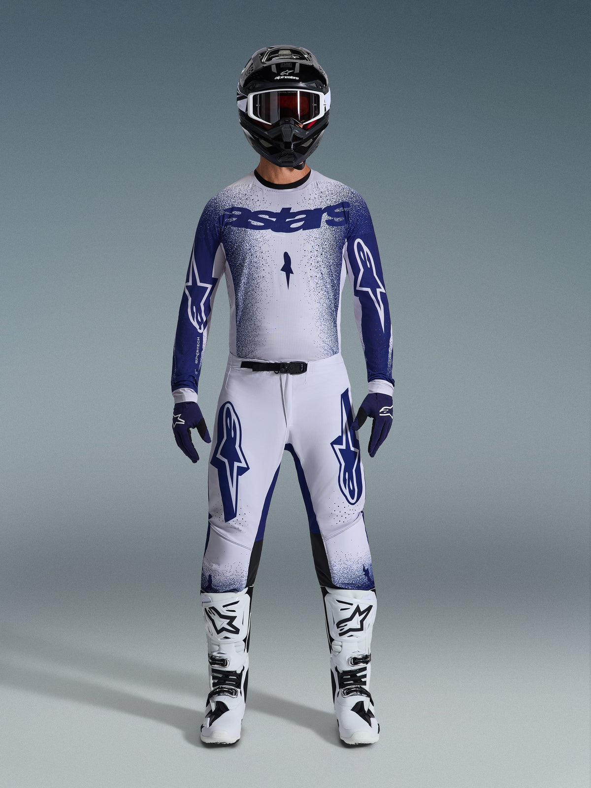 Motocross-Fahrer im Alpinestars 2026 Supertech Scenz Gear Set, Hellgrau und Dunkelblau, inklusive Trikot und Hose mit passenden blauen Handschuhen, weiß-schwarzen Stiefeln und einem schwarzen Helm mit weißer Brille, koordiniertes Outfit für professionellen Offroad-Rennsport