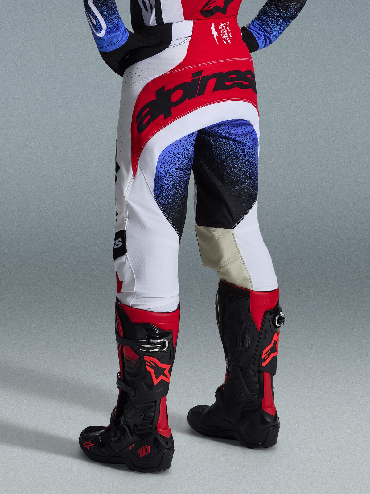 Alpinestars 2026 Techstar Knif Hose, Motocross-Hose, Schwarz, Weiß und Rot mit blauen Akzenten, Rückansicht mit markantem Branding am Bund und hellbraunen Leder-Innenknie-Einsätzen, getragen mit schwarz-roten Stiefeln, Hochleistungs-Rennausrüstung für Offroad-Wettbewerbe