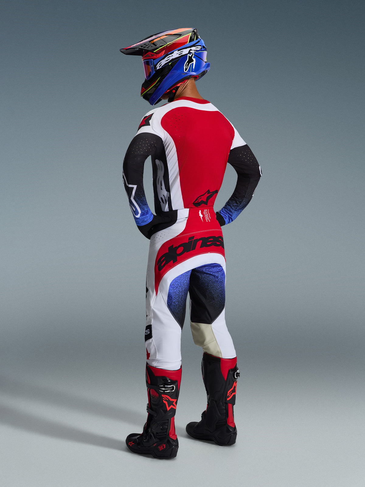 Alpinestars 2026 Techstar Knif Hose, Motocross-Hose, Schwarz, Weiß und Rot mit blauen Farbverlauf-Akzenten, Rückansicht eines Fahrers in kompletter Rennausrüstung, markantes rotes Branding auf der Sitzfläche, weiße und schwarze Einsätze, getragen mit passendem Jersey und Tech 10 Stiefeln.