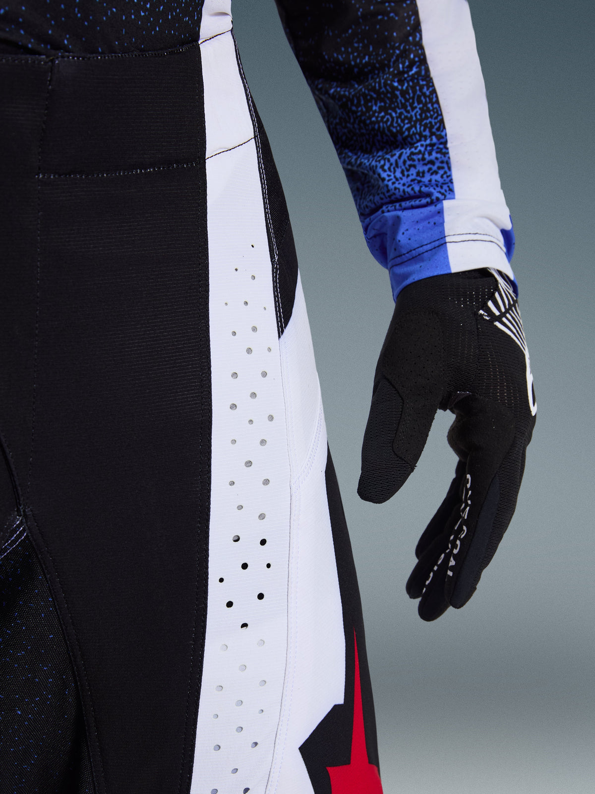 Alpinestars 2026 Techstar Knif Hose, Motocross-Hose, Schwarz, Weiß und Rot, Nahaufnahme der lasergeschnittenen Perforationen an den Seitenbahnen und des roten Stern-Logos, kombiniert mit einem passenden Trikot und schwarzem Motocross-Handschuh.