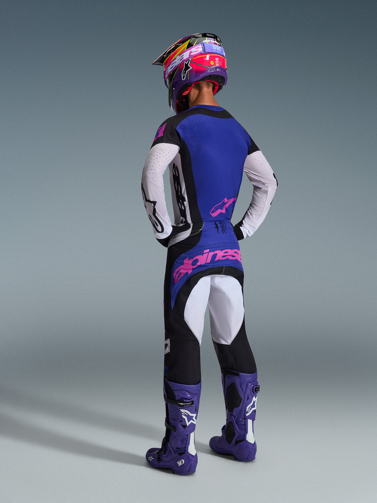 Motocross rider wearing the 2026 Techstar Knif Gear Set from a rear view, featuring a blue and white jersey with pink accents, black pants with light gray panels and bold pink branding, purple racing boots, and a matching purple and pink helmet, coordinated off-road racing gear.
