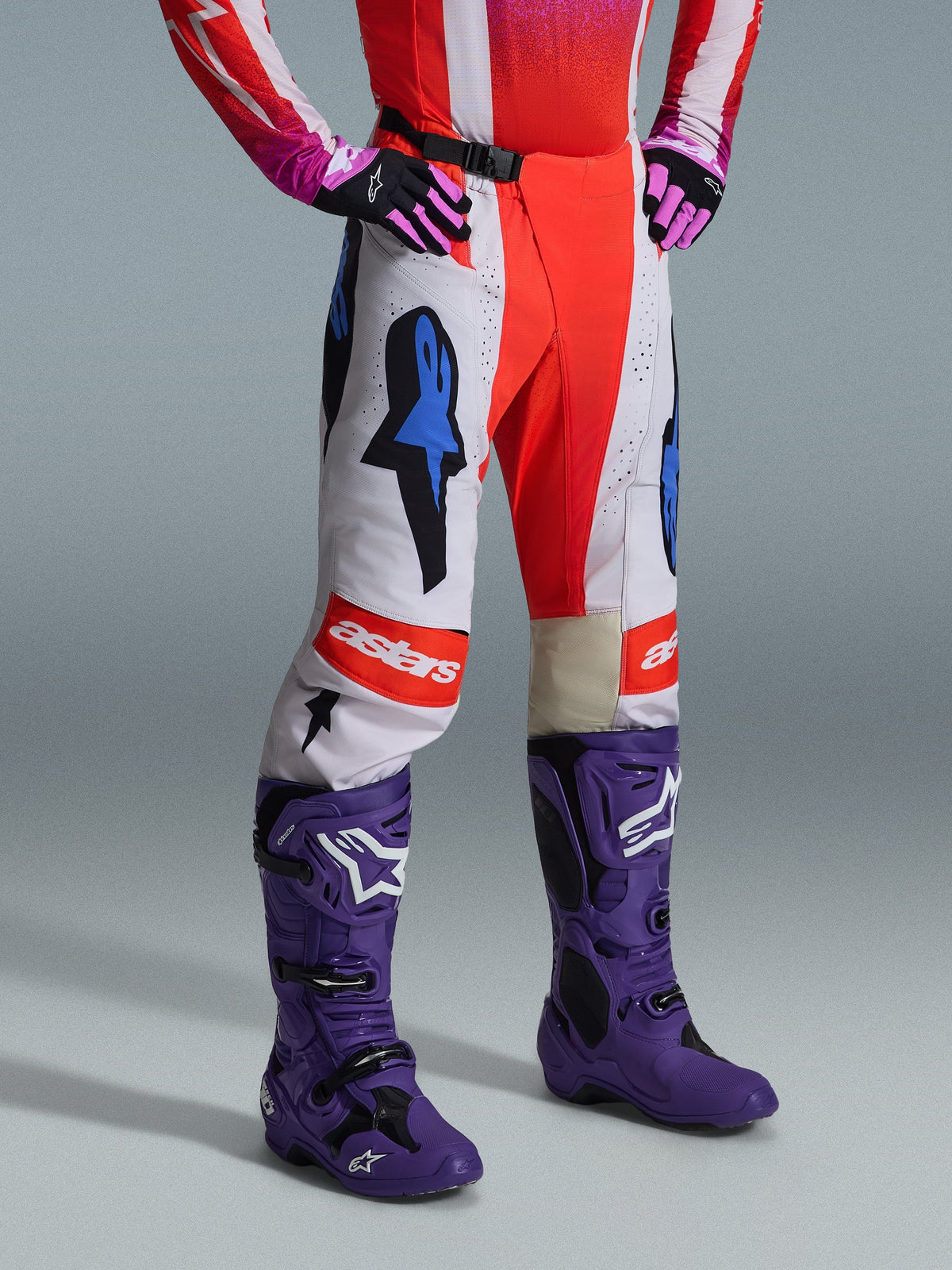 Alpinestars 2026 Techstar Knif Hose, Motocross-Hose, Weiß und Orange mit blauen Akzenten, getragen als Teil einer kompletten Ausrüstung mit einem rot-pinken Jersey, lila Stiefeln und pinken Handschuhen, mit markanten Logos und einer athletischen Rennpassform