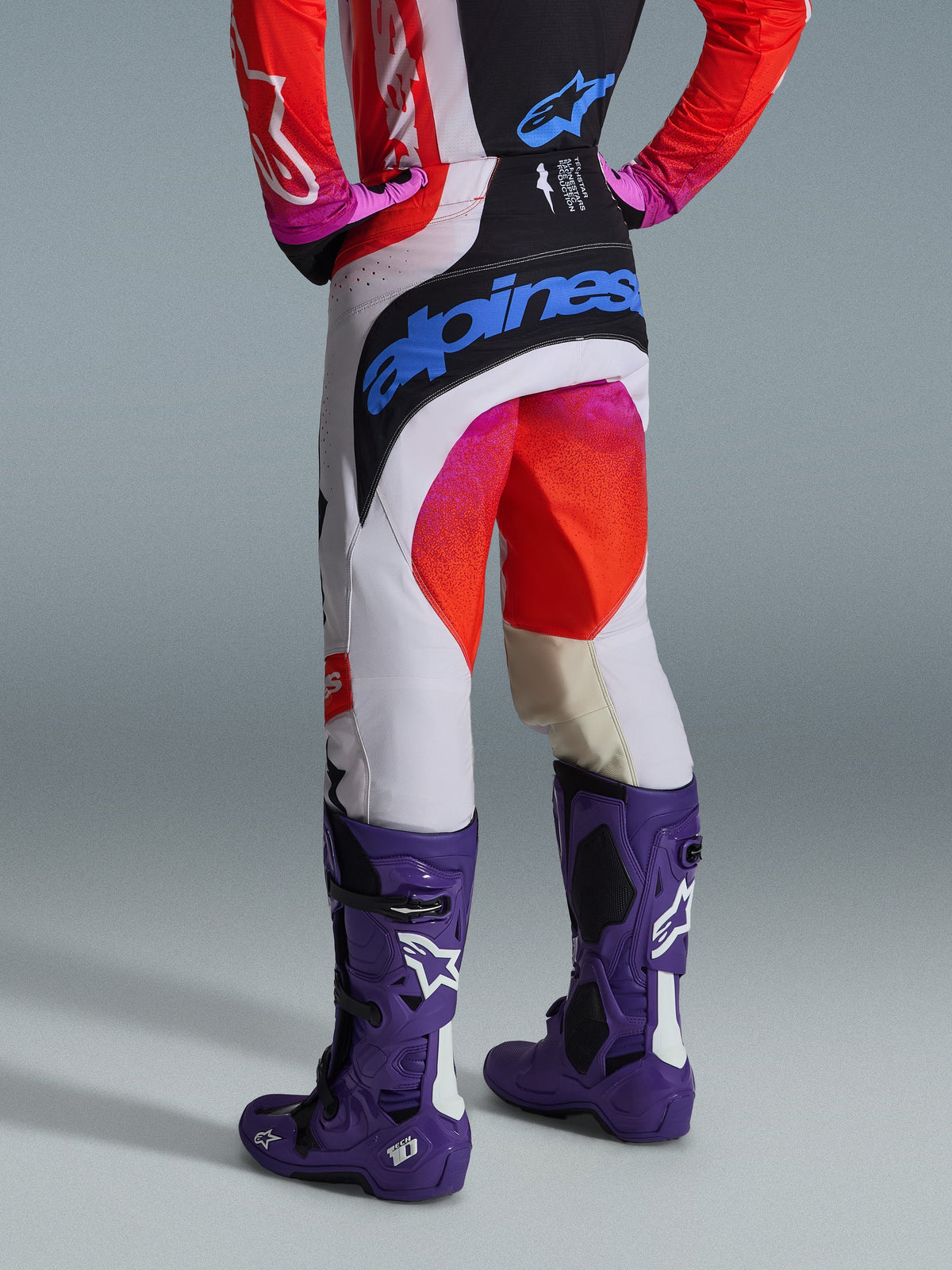 Alpinestars 2026 Techstar Knif Hose, Motocross-Hose, Lichtgrau, Orange Fluo und Schwarz, Rückansicht mit großem blauem Alpinestars-Branding am Gesäß, kombiniert mit violetten Tech 10 Stiefeln, professionelle Offroad-Rennausrüstung