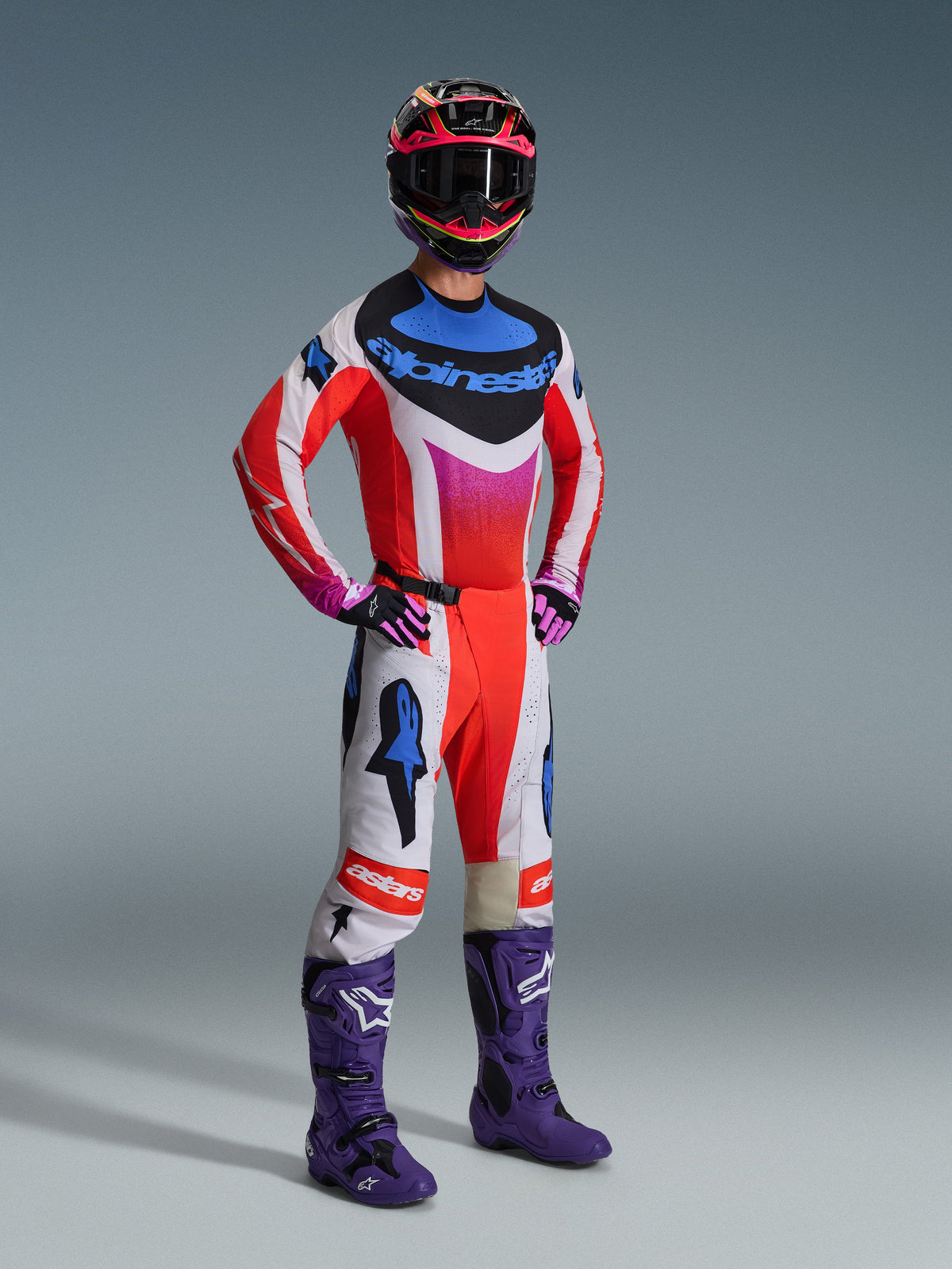 Alpinestars 2026 Techstar Knif Gear Set, Motocross-Fahrer in voller Rennausrüstung, rotes Jersey mit weißen und blauen Akzenten, weiße Hose mit roten und blauen Stern-Logos, lila Motocross-Stiefel, schwarz-pinker Helm, professionelles Offroad-Rennset