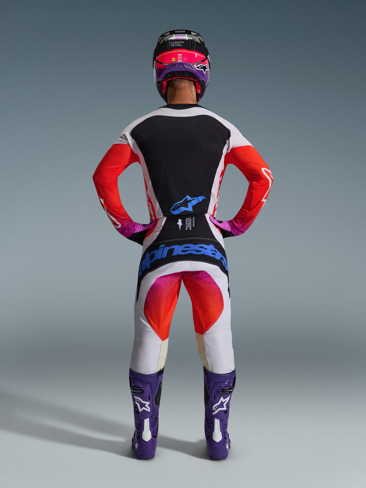 Motocross-Fahrer in der Rückansicht mit 2026 Techstar Knif Hose und passendem Trikot, hellgraues und schwarzes Design mit leuchtend orangefarbenen und lila Akzenten, blaues Alpinestars-Logo auf der Gesäßpartie, lila Stiefel und Helm, professionelles Offroad-Rennausrüstungsset.