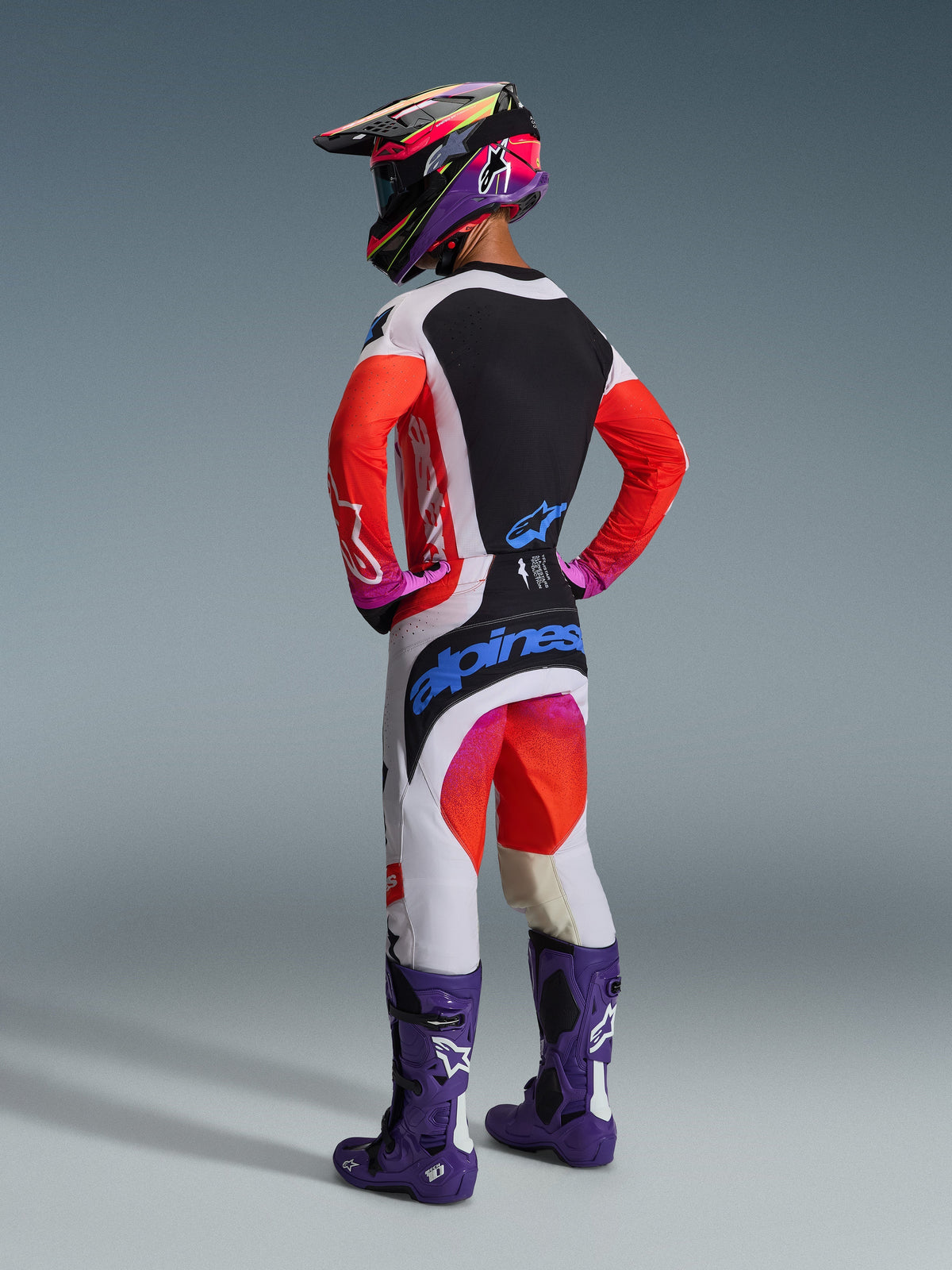 Motocross-Fahrer in der Rückansicht in kompletter Rennausrüstung, Alpinestars 2026 Techstar Knif Hose in Hellgrau und Orange Fluo mit blauem Branding, passendes Jersey mit orangefarbenen Ärmeln, lila Tech 10 Stiefel und ein farbenfroher Rennhelm, professionelles Offroad-Outfit