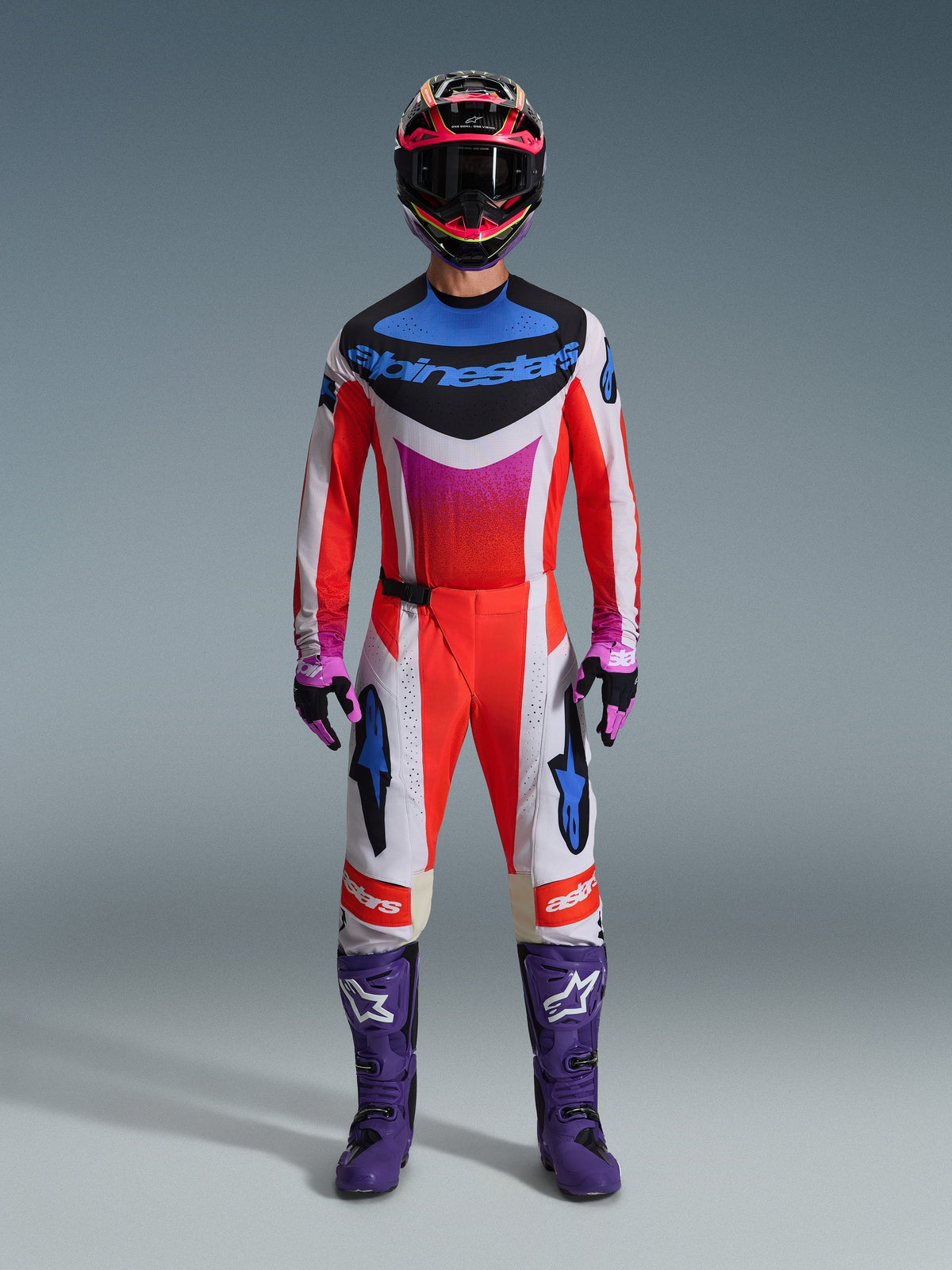 Motocross-Fahrer in kompletter Rennausrüstung, Alpinestars 2026 Techstar Knif Hose, fluo-orangefarbenes und weißes Design mit blauen Akzenten, passendes mehrfarbiges Trikot, lilafarbene Stiefel, schwarz-lila Handschuhe, koordiniertes Profi-Outfit für Motocross-Rennen