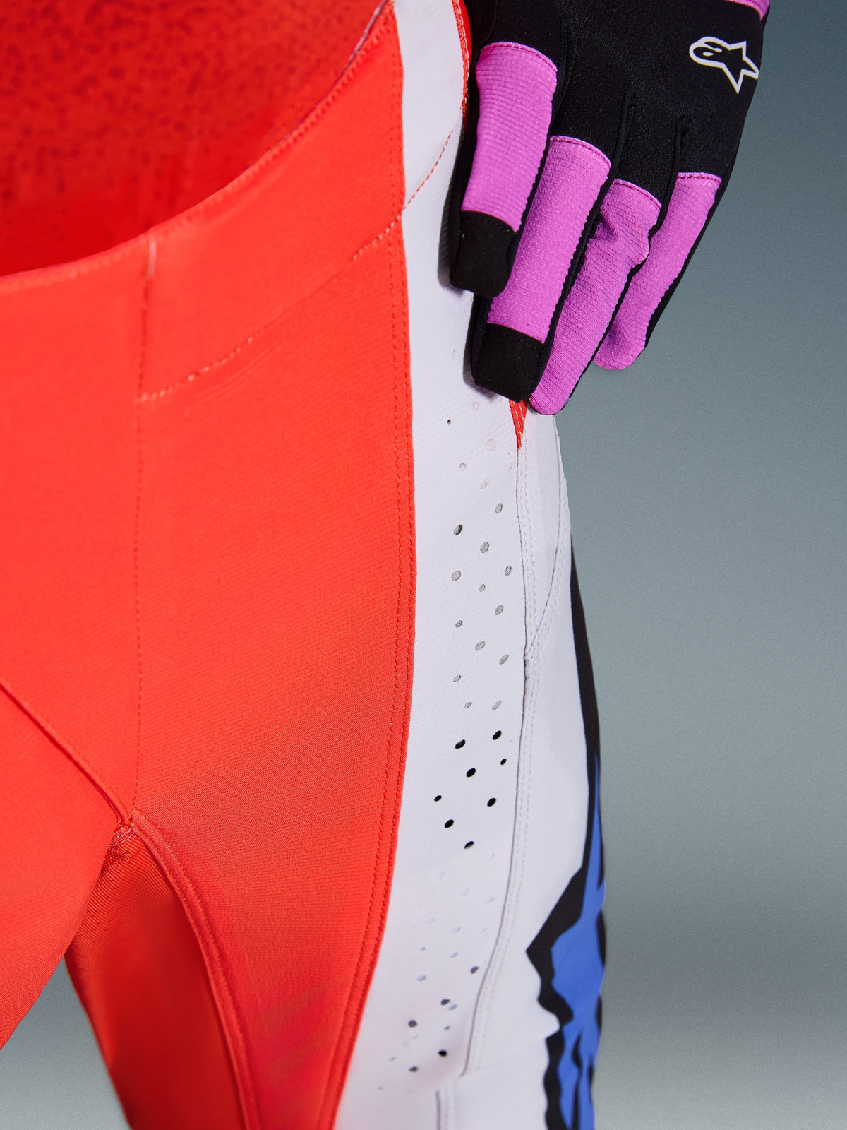 Alpinestars 2026 Techstar Knif Hose, Nahaufnahme der Hüfte und des Seitenpanels in Orange Fluo und Hellgrau, mit lasergeschnittenen Perforationen für Luftzirkulation, kombiniert mit einem schwarz-lila Motocross-Handschuh, Hochleistungs-Rennausrüstung