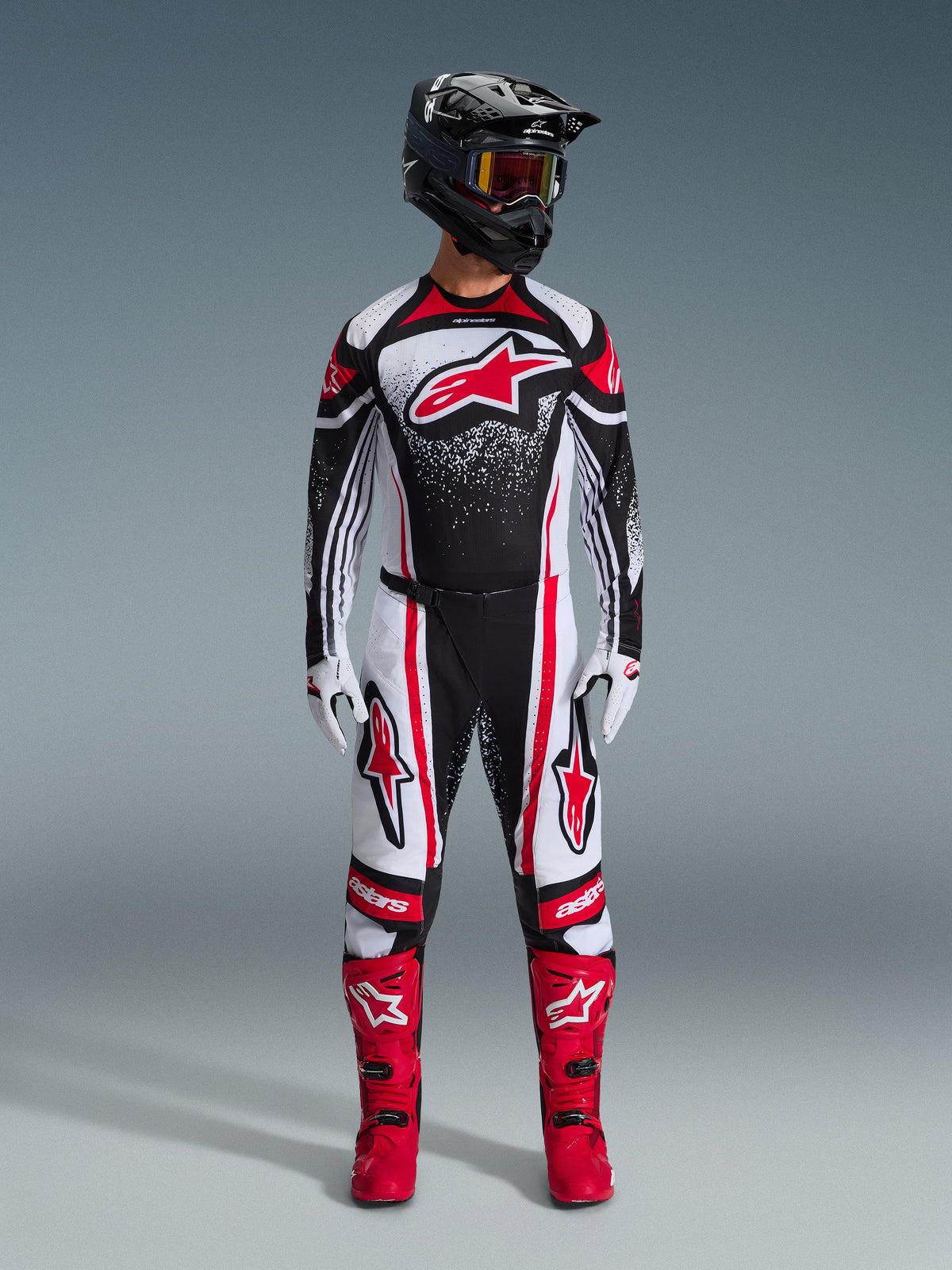 Alpinestars 2026 Techstar Nomur Gear Set, Motocross rider standing in full racing gear, Black white and red design with white speckle pattern, jersey featuring red Astars logo, matching white and red pants, bright red boots, and black helmet with goggles, coordinated high-performance motocross kit