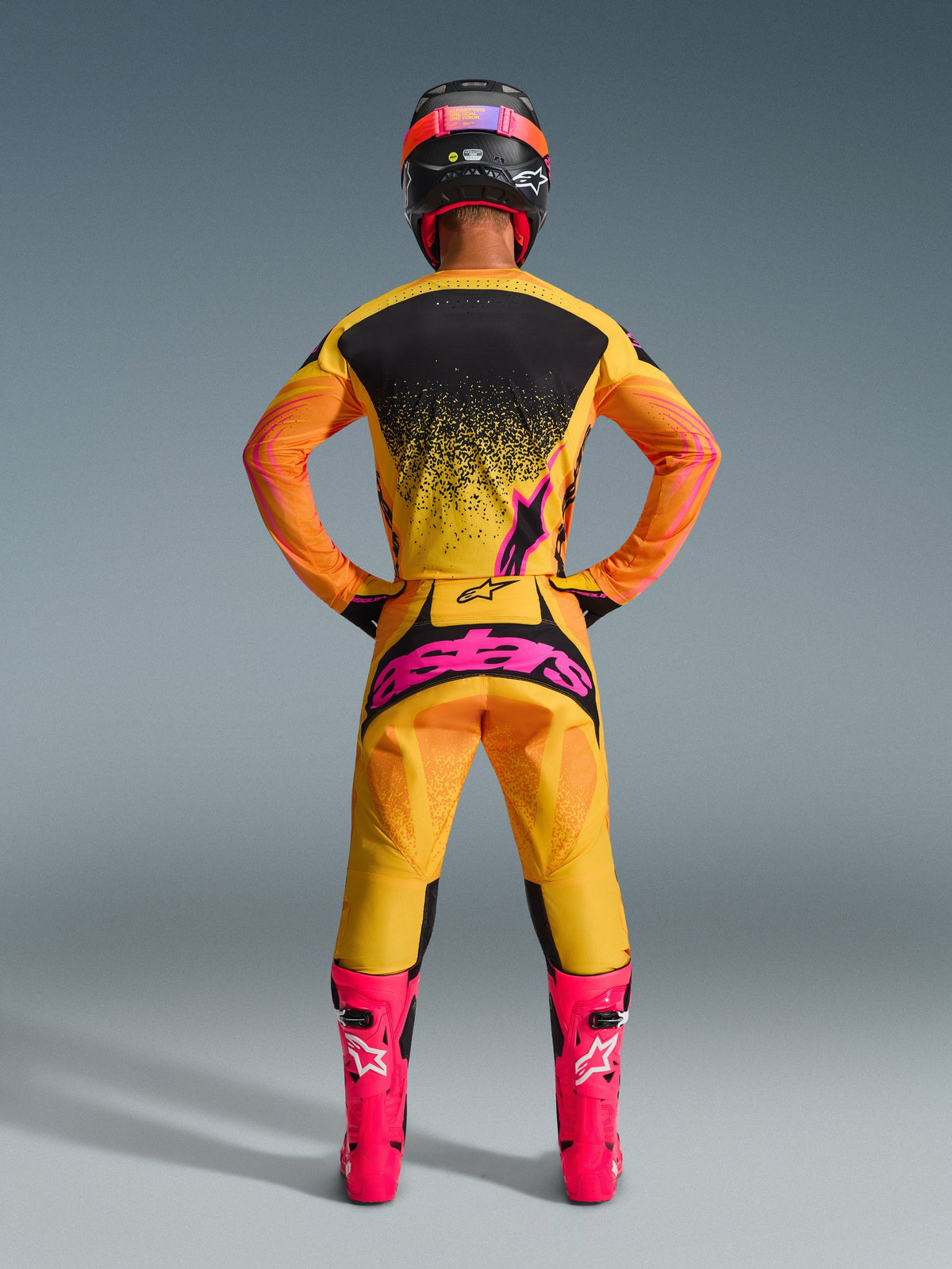Rear view of a motocross rider wearing the Alpinestars 2026 Techstar Nomur Pants in Orange Yellow Pink, featuring a large pink logo on the seat, paired with a matching yellow and black jersey, bright pink boots, and a black and pink helmet.