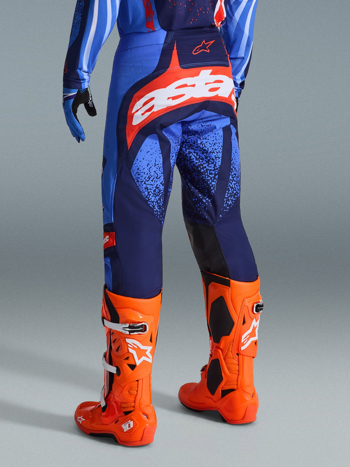 Alpinestars 2026 Techstar Nomur Hose, Motocross-Hose, Dunkelblau, Orange und UCLA-Blau, Rückansicht mit weißem „Astars“-Logo auf dem Gesäß, getragen mit leuchtend orangen Motocross-Stiefeln und passendem Trikot, athletische Rennpassform für Offroad-Wettkämpfe.