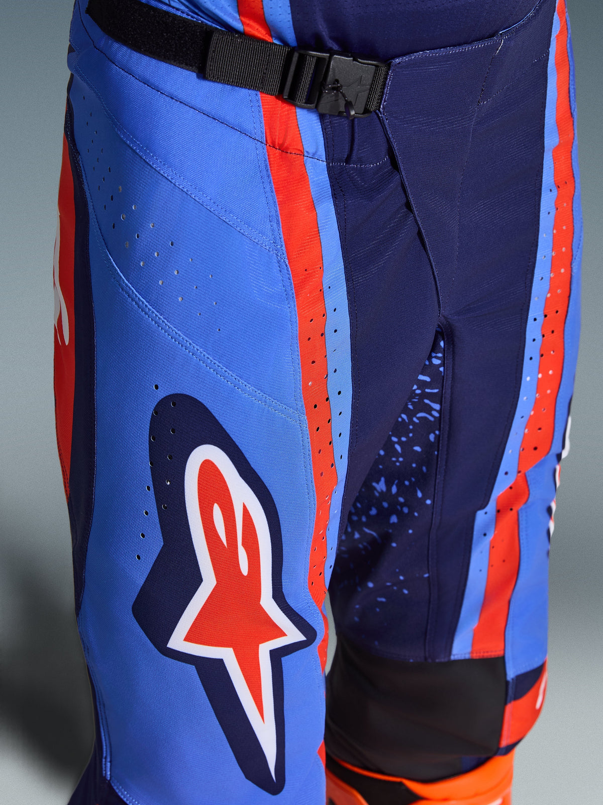 Alpinestars 2026 Techstar Nomur Hose, Motocross-Hose in Dark Navy Orange und UCLA Blue, Nahaufnahme des SX2-Magnetverschlusses am Bund, lasergeschnittene Perforationen am Oberschenkel, ikonisches Stern-Logo, strapazierfähige Offroad-Rennausrüstung.