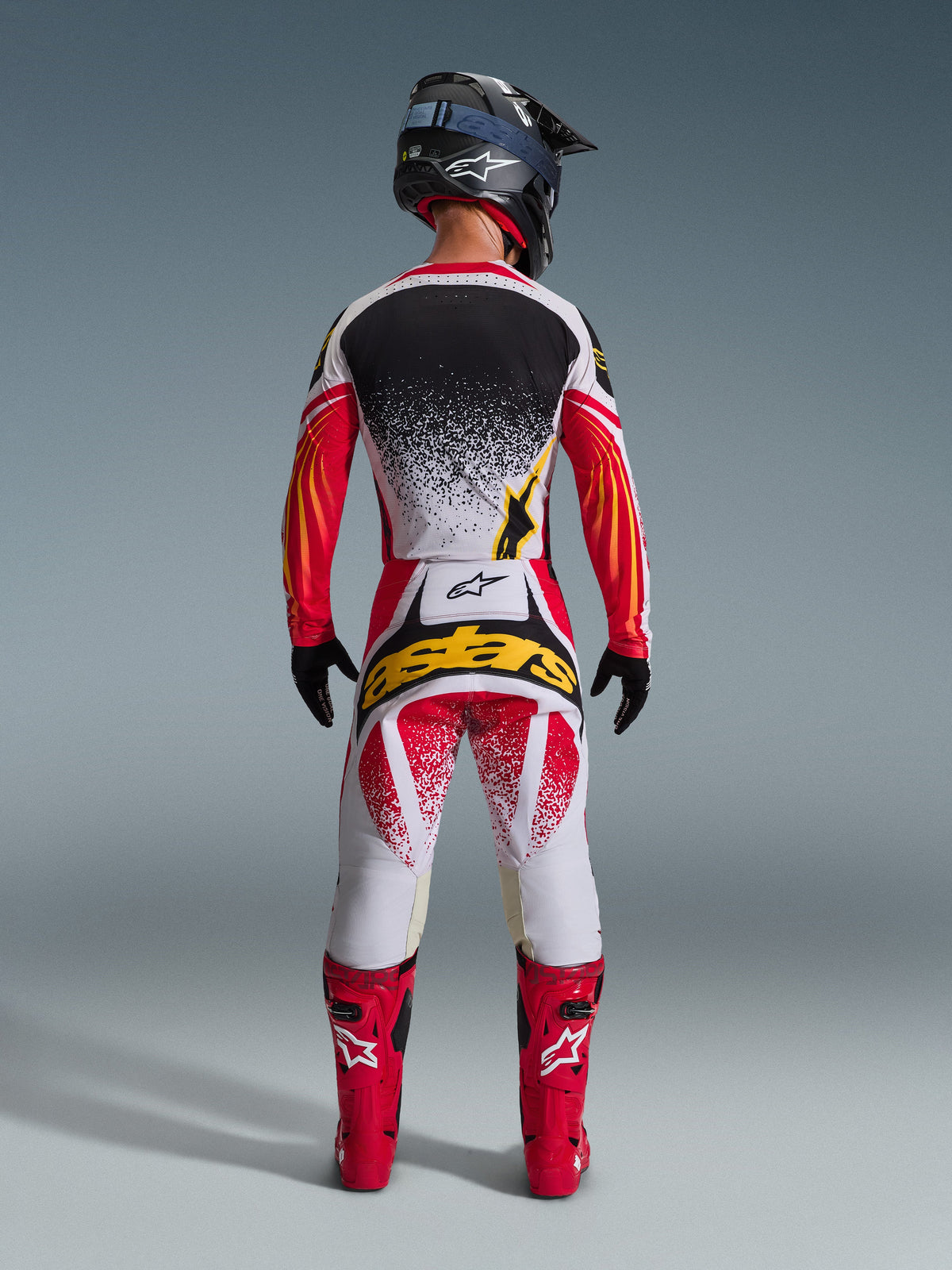 Alpinestars 2026 Techstar Nomur Gear Set, komplette Motocross-Rennausrüstung in der Rückansicht, weißes Jersey mit schwarzem Splatter-Effekt und rot-orangen Grafik-Ärmeln, weiß-rote Hose mit gelbem 'astars'-Logo, rote Rennstiefel, schwarzer Helm in Carbon-Optik mit blauer Brille, Hochleistungs-Offroad-Ausrüstung.