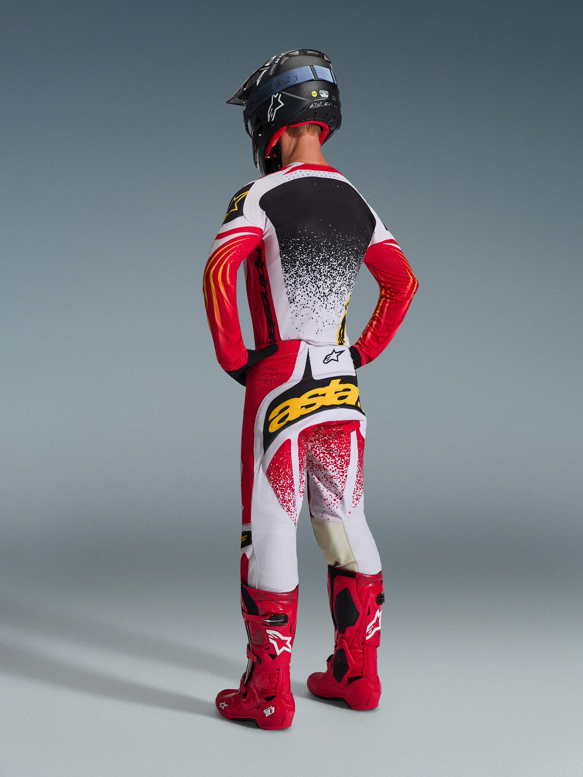 Motocross-Fahrer von hinten im Alpinestars 2026 Techstar Nomur Gear Set, rot-weiß-schwarzes Trikot mit weiß gesprenkeltem Rücken, passende Hose mit gelbem 'astars'-Logo auf dem Gesäß, leuchtend rote Rennstiefel und schwarzer Helm, komplette koordinierte Motocross-Ausrüstung