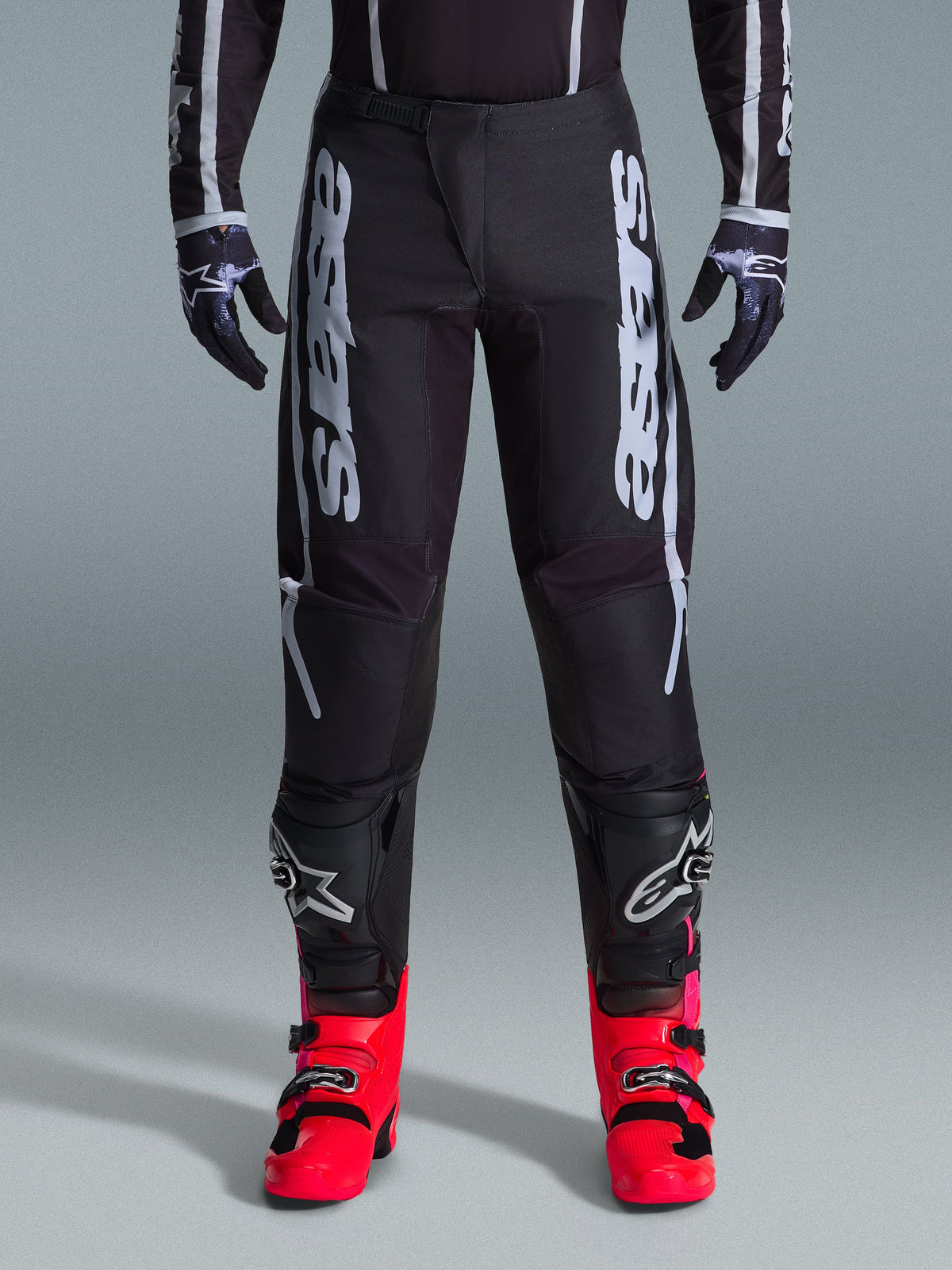 Alpinestars 2026 Fluid Apex Hose, Motocross-Hose in Schwarz mit großer weißer Beschriftung, getragen mit schwarzem Knieschutz und leuchtend roten Stiefeln, leichtes 600D-Polygewebe, Sport-Fit-Design für den Offroad-Rennsport
