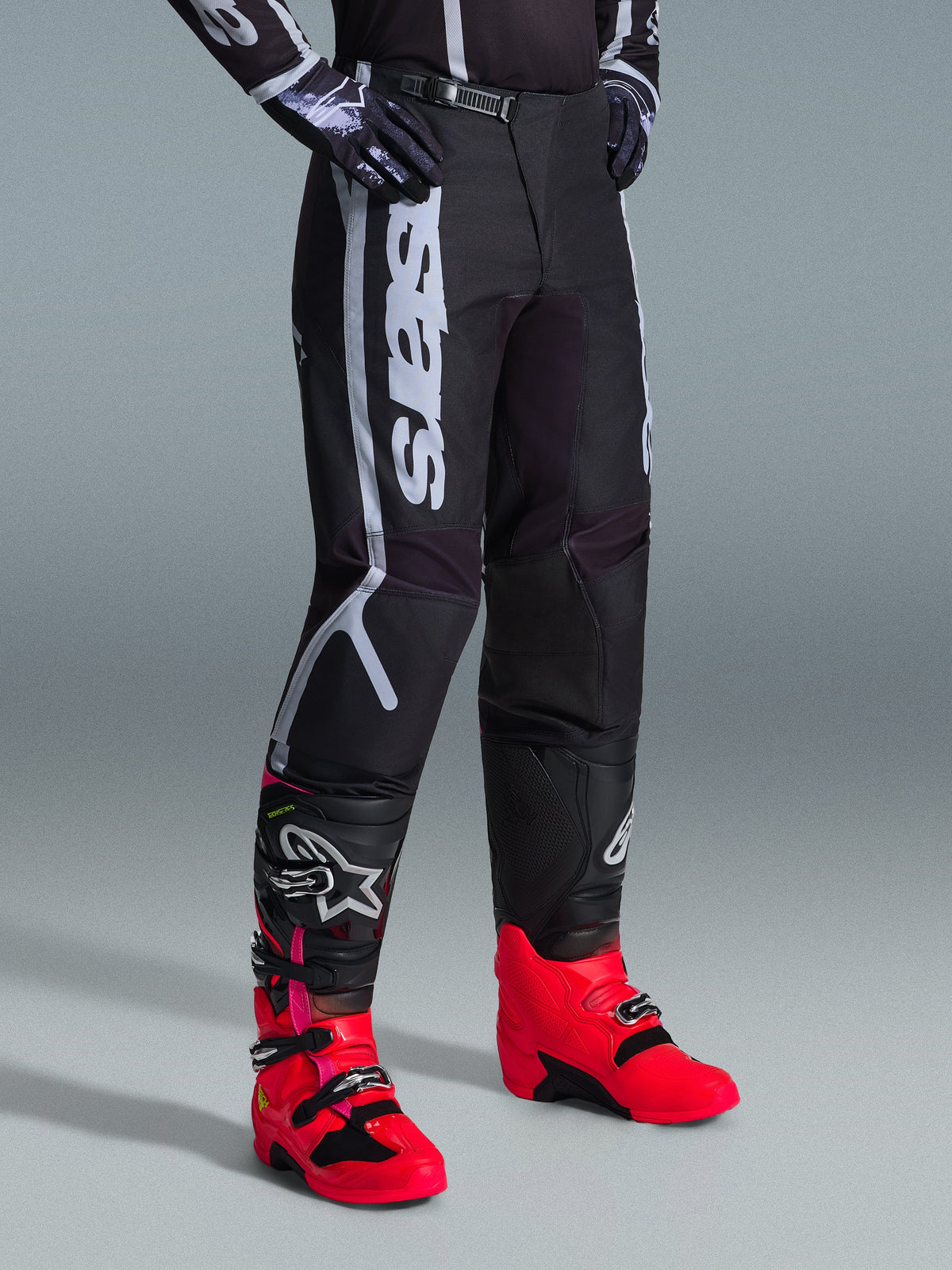 Alpinestars 2026 Fluid Apex Hose, Motocross-Hose, Schwarz und Grau, getragen mit leuchtend pinkfarbenen und roten Stiefeln sowie passendem schwarz-weißem Trikot, mit markantem vertikalem Alpinestars-Schriftzug am Oberschenkel, verstärkten Einsätzen und Ratschenverschluss für optimale Offroad-Performance.