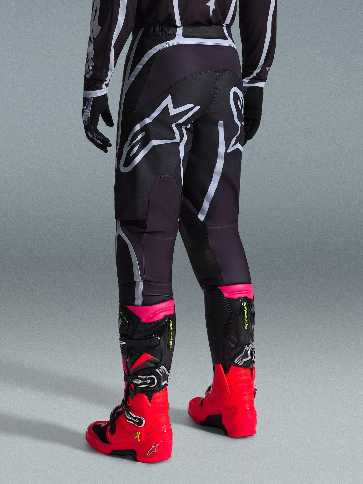 Alpinestars 2026 Fluid Apex Hose, Motocross-Hose, Schwarz und Grau, Rückansicht mit großen weißen Alpinestars-Logos am Gesäß, kombiniert mit leuchtend roten und pinken Stiefeln, strapazierfähige 600D-Polygewebe-Konstruktion für Offroad-Performance