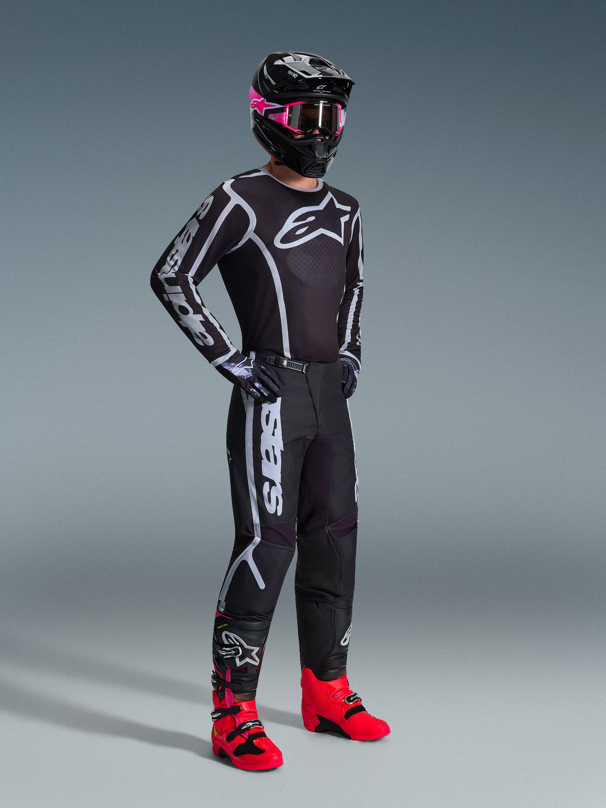 Alpinestars 2026 Fluid Apex Hose, Motocross-Hose, Schwarz und Grau, kombiniert mit passendem schwarz-weißem Trikot, schwarzem Helm mit pinker Brille und leuchtend roten Stiefeln, mit auffälligem weißem Branding an den Beinen und einem sportlichen Design für den Offroad-Rennsport.