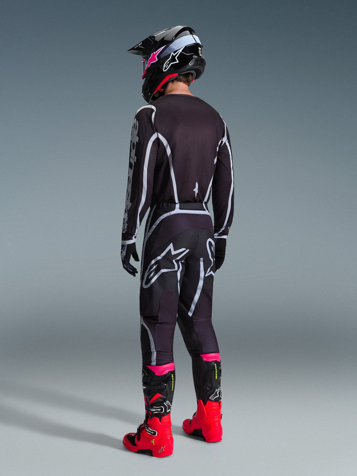 Alpinestars 2026 Fluid Apex Hose, Motocross-Hose, Schwarz und Grau, Rückansicht mit weißem Alpinestars-Logo auf dem Gesäß, getragen mit passendem Trikot, schwarz-pinkem Helm und leuchtend roten Stiefeln.