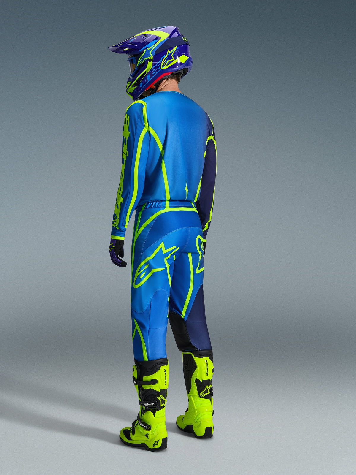 Motocross rider from a rear view wearing 2026 Alpinestars Fluid Apex Pants in Blue and Yellow Fluo, matching blue and yellow jersey and helmet, and bright neon yellow boots, coordinated racing gear for off-road performance.
