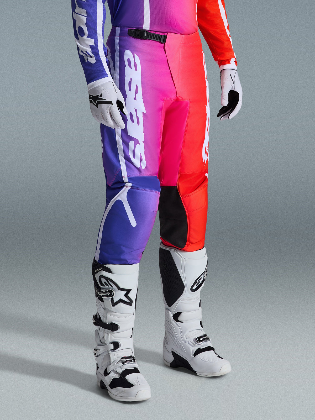 Alpinestars 2026 Fluid Apex Hose, Motocross-Hose, mehrfarbiger Farbverlauf in Lila, Pink und Rot, getragen mit weiß-schwarzen Motocross-Stiefeln und Handschuhen, mit weißem vertikalem Branding und schwarzen Knie-Akzenten, Performance-Offroad-Ausrüstung