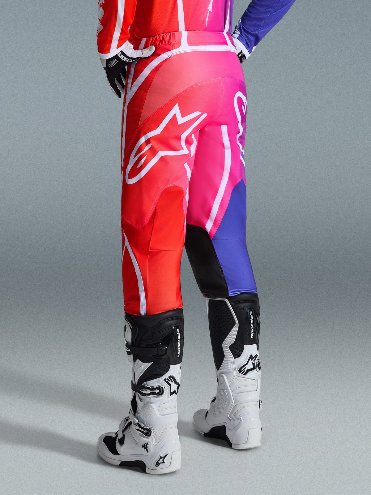 Alpinestars 2026 Fluid Apex Hose, Motocross-Hose, Mehrfarbig in Rot, Pink und Lila, Rückansicht eines Fahrers mit weiß-schwarzen Motocross-Stiefeln, großes weißes Alpinestars-Logo, lebendiges Farbverlaufs-Design