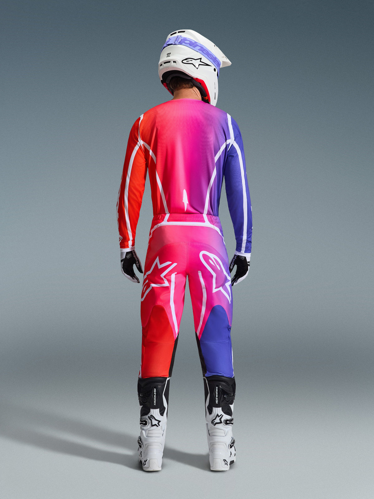 Rückansicht eines Motocross-Fahrers mit Alpinestars 2026 Fluid Apex Hose und passendem Trikot, lebendiger Farbverlauf in Rot, Pink und Lila, weißer Helm und weiße Stiefel, komplettes abgestimmtes Ausrüstungsset für den Offroad-Rennsport