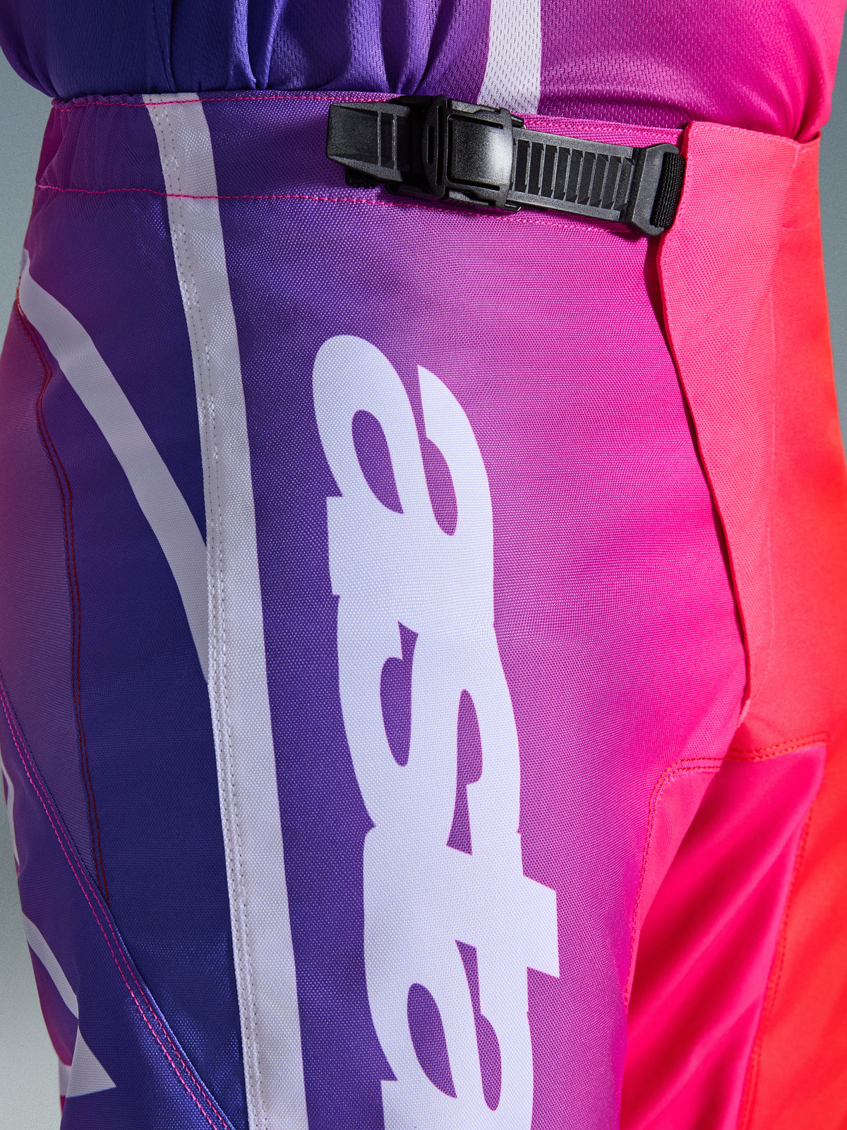 Alpinestars 2026 Fluid Apex Hose, Motocross-Hose, Mehrfarbig Lila, Pink und Rot, Nahaufnahme des Taillen- und Hüftbereichs mit schwarzem Ratschenverschluss-System und großem weißen vertikalen Alpinestars-Logo, lebendiges Farbverlauf-Design mit strapazierfähiger Polygewebe-Struktur