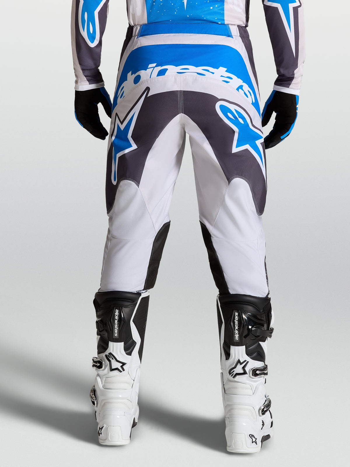 Alpinestars Fluid Drag Hosen, Herren-Motocross-Hose in Grau und Blau, verstärkter Sitzbereich für Offroad-Fahrten, Rückansicht am Fahrer mit weißen Motorradstiefeln.