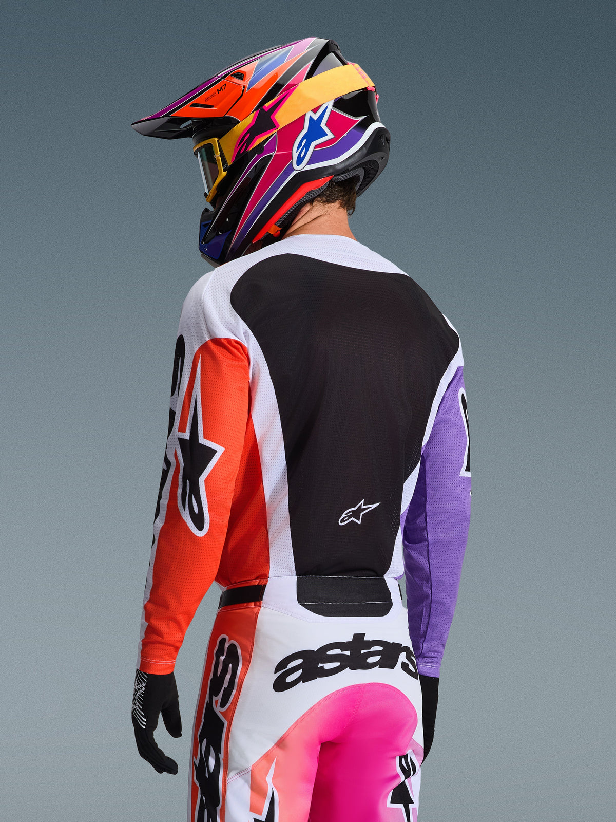 Motocross-Fahrer in der Rückansicht mit dem Alpinestars 2026 Racer Air Portl Trikot in Multicolor, orangefarbener rechter Ärmel, lila linker Ärmel, großes schwarzes Mesh-Rückenteil zur Belüftung, passende Motocross-Hose und farbenfroher Helm, professionelle Offroad-Rennausrüstung.