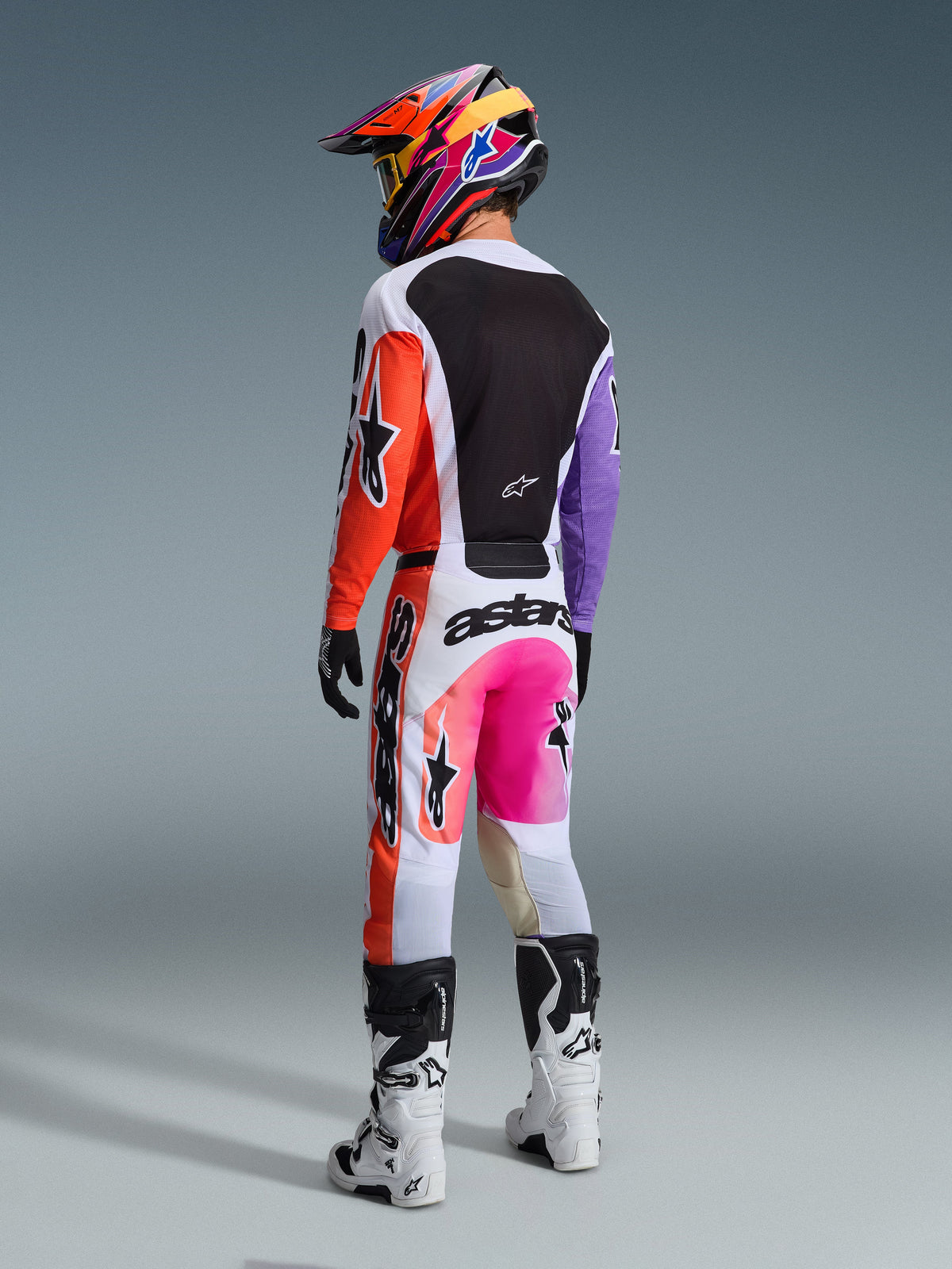 Alpinestars 2026 Racer Air Portl Trikot, Mehrfarbiges Motocross-Trikot, Rückansicht eines Fahrers in voller Rennausrüstung, mit weißer Basis, orangefarbenen und lila Ärmeln sowie schwarzem Mesh-Rückeneinsatz, kombiniert mit passender pinker und orangefarbener Hose, buntem Helm und weißen Stiefeln, professionelles Offroad-Rennoutfit