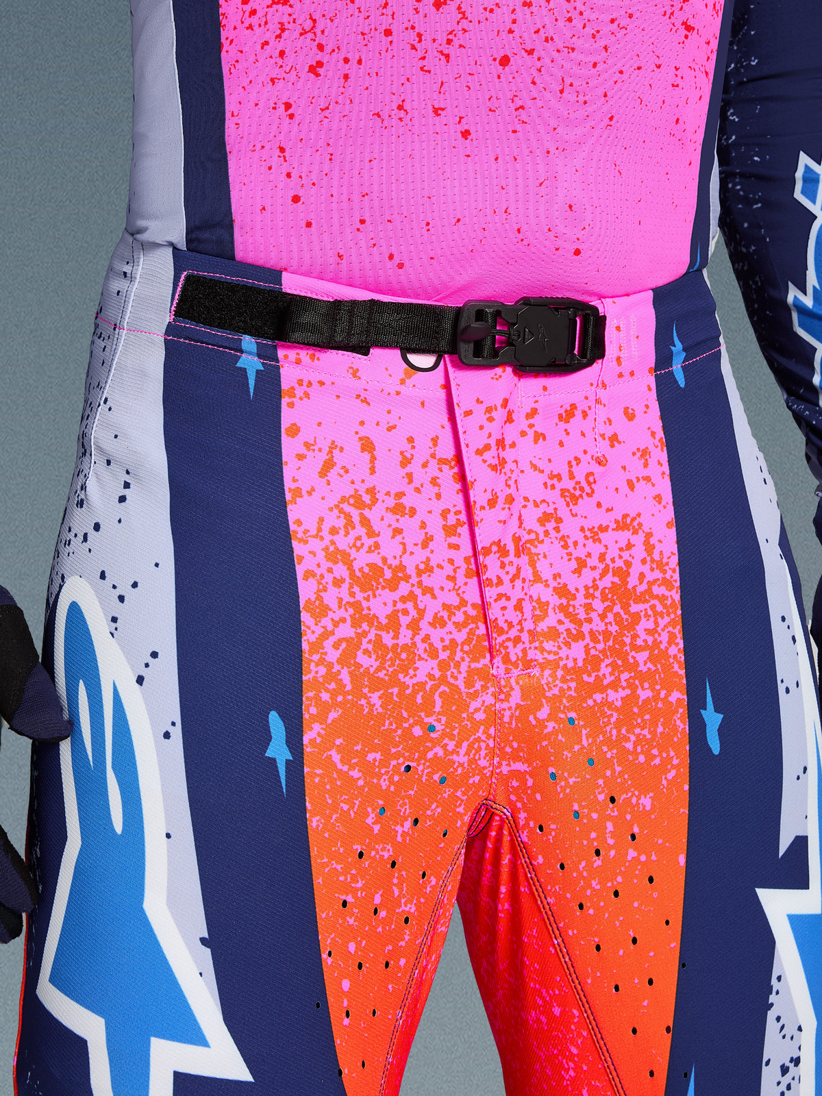 Alpinestars Limited Edition Supertech Records Pants, Motocross Pants, Multicolor pink and orange with navy blue side panels, close-up view of the waist featuring a black SX1 auto-locking magnetic buckle, rip-stop fabric with splatter graphics and laser-cut perforations.
