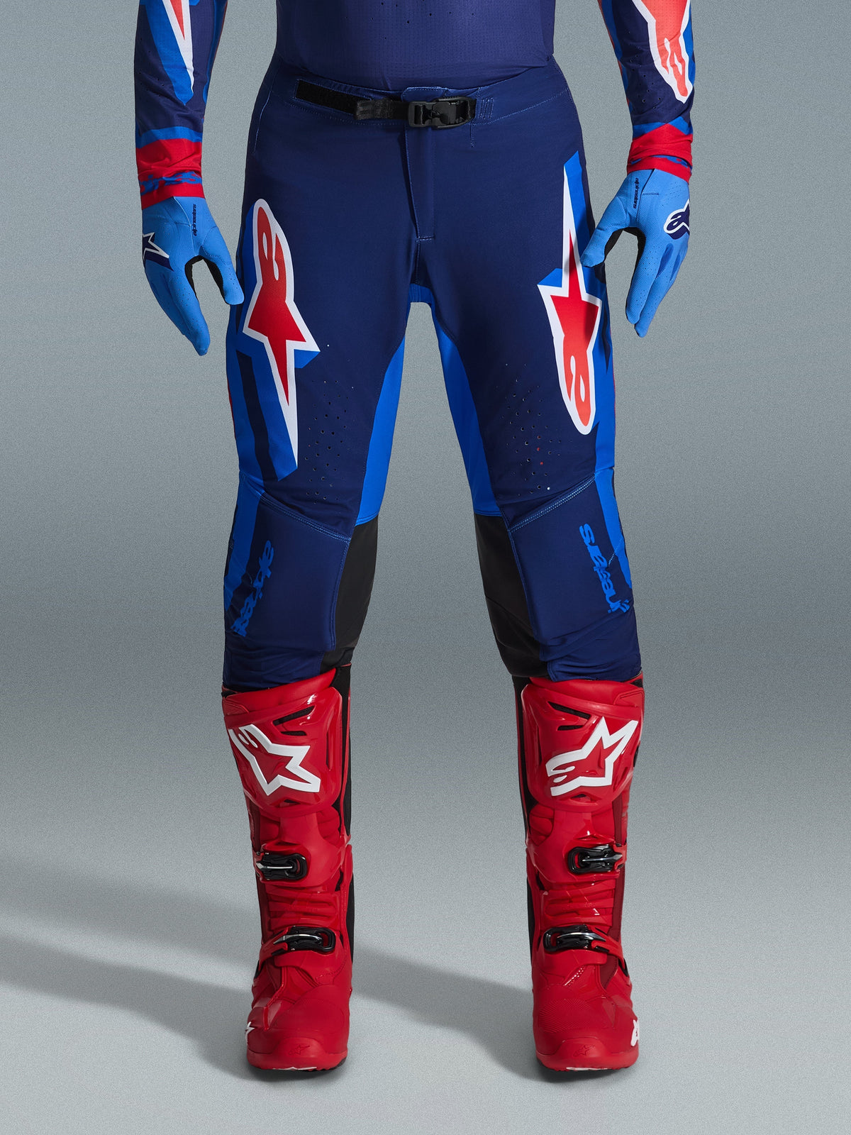 Alpinestars 2026 Supertech Vista Hose, Motocross-Hose, Dark Navy, Hot Coral und Cobalt, Frontansicht eines Fahrers in Performance-Ausrüstung mit passenden blauen Handschuhen und roten Motocross-Stiefeln, mit markanten roten und weißen Alpinestars-Logos auf den Oberschenkeln.