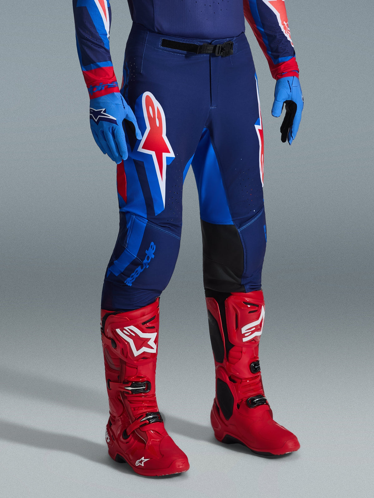 Alpinestars 2026 Supertech Vista Hose, Motocross-Hose, Dunkelblau, Hot Coral und Kobaltblau, getragen mit leuchtend roten Rennstiefeln und blauen Handschuhen, mit großen grafischen Logos auf den Oberschenkeln und schwarzen verstärkten Knie-Innenpaneelen für Offroad-Performance.