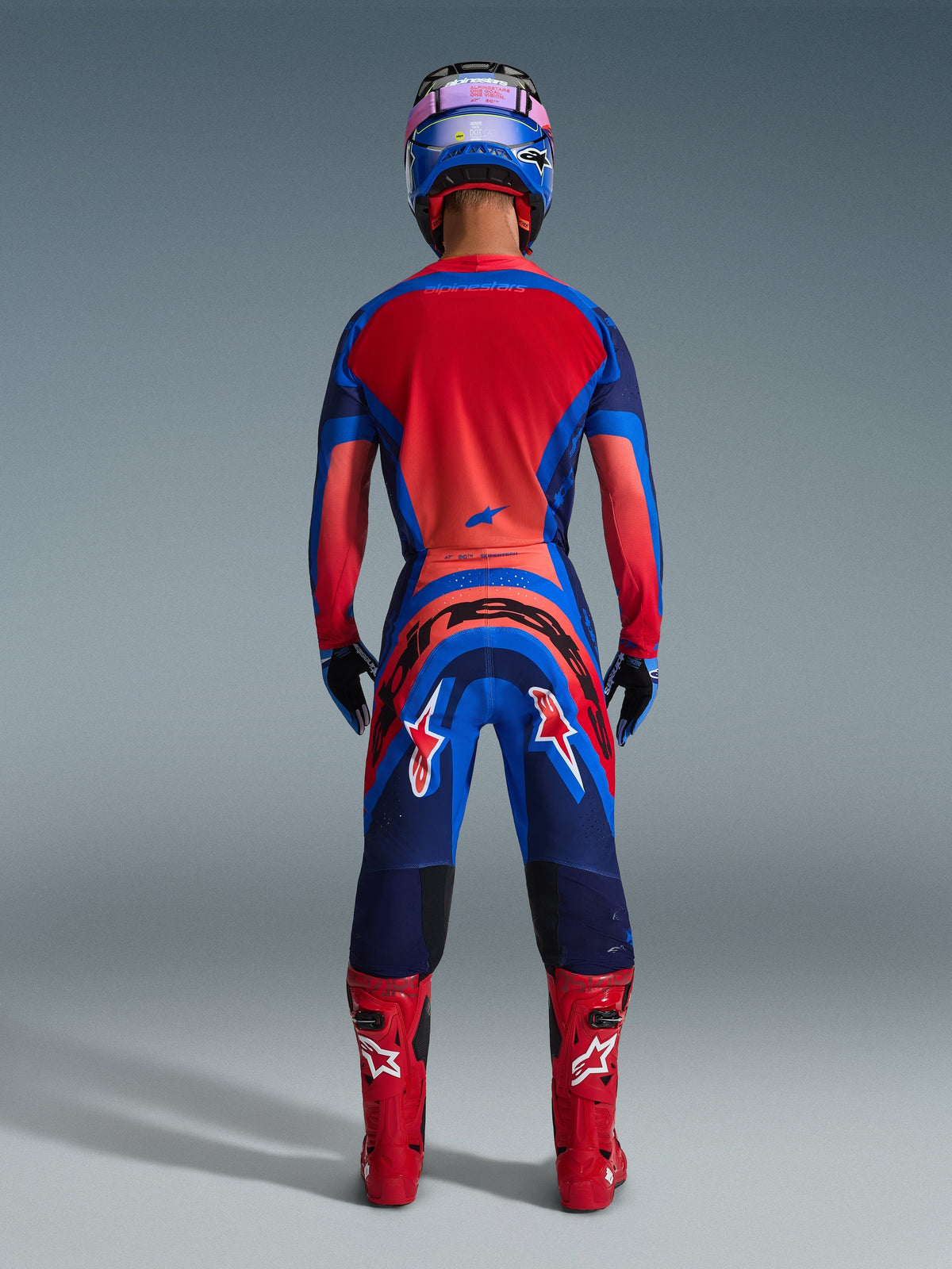 Alpinestars 2026 Supertech Vista Hose, Motocross-Hose, Dark Navy, Hot Coral und Cobalt, Rückansicht eines Fahrers in kompletter Rennausrüstung mit passendem Trikot, blauem Helm und roten Stiefeln, mit auffälligem Branding auf dem Gesäß und den Beinen.