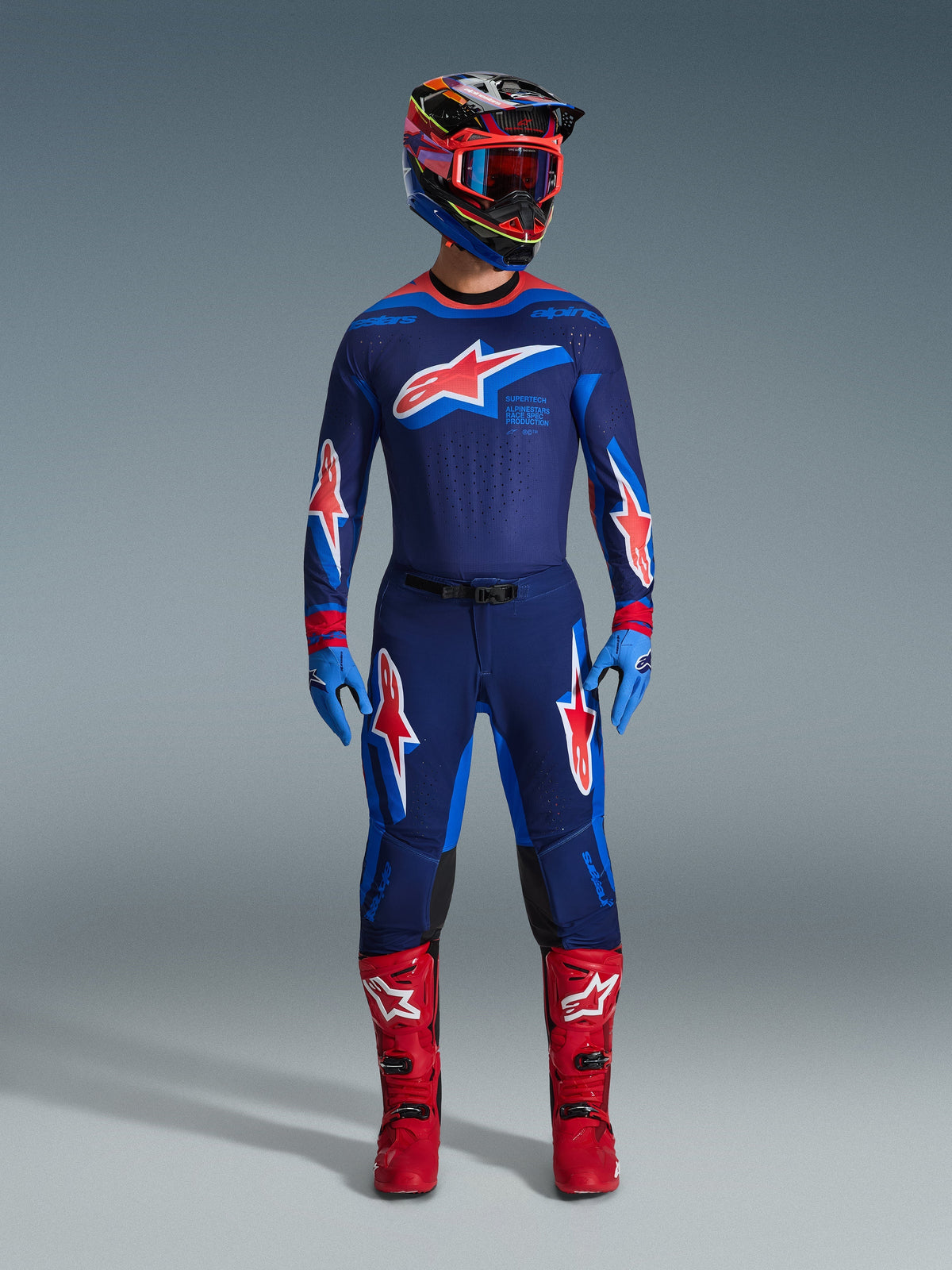 Motocross-Fahrer in vollständiger Rennausrüstung, Alpinestars 2026 Supertech Vista Hose in Dark Navy Hot Coral Cobalt, passendes Trikot mit markantem Stern-Logo, leuchtend rote Stiefel und Helm mit roter Brille, abgestimmtes Profi-Set für Offroad-Performance