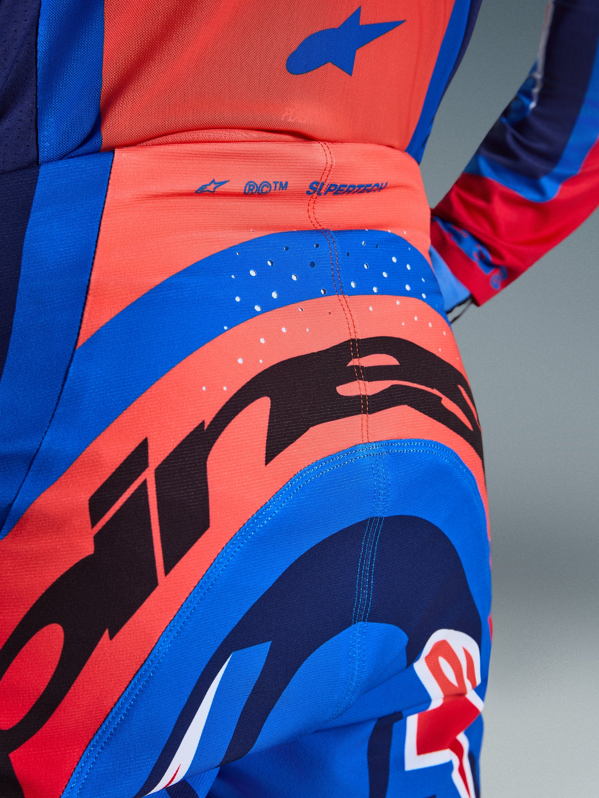 Nahaufnahme der Alpinestars 2026 Supertech Vista Hose, Motocross-Hose, Hot Coral und Cobalt Blue, mit einer großen schwarzen Alpinestars-Grafik am Gesäß, lasergeschnittenen Belüftungslöchern und „SUPERTECH“-Branding am hinteren Bund, professionelle Rennbekleidung.