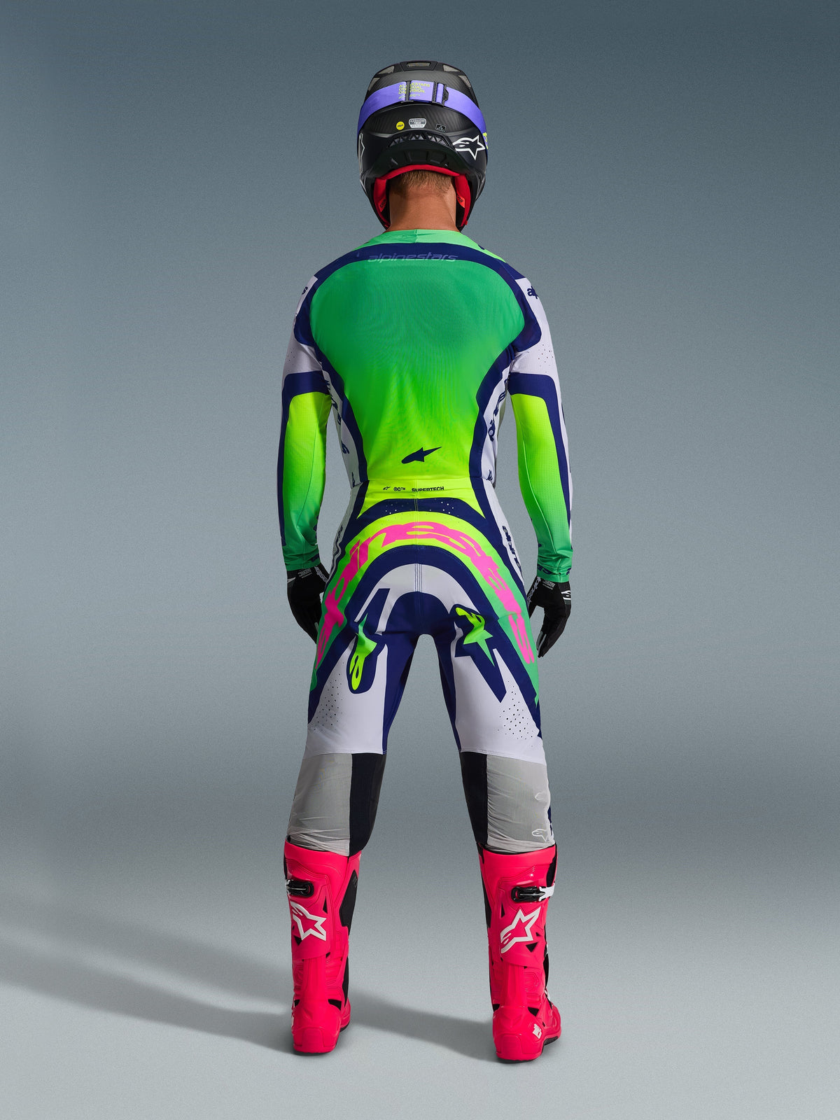 Alpinestars 2026 Supertech Vista Hose, Motocross-Hose, Hellgrau, Gelb Fluo und Blau, Rückansicht eines Fahrers in einem passenden grün-gelben Trikot mit Farbverlauf und leuchtend pinken Stiefeln, mit markantem pinken Alpinestars-Branding auf dem Gesäß, professionelle athletische Passform für den Offroad-Rennsport.