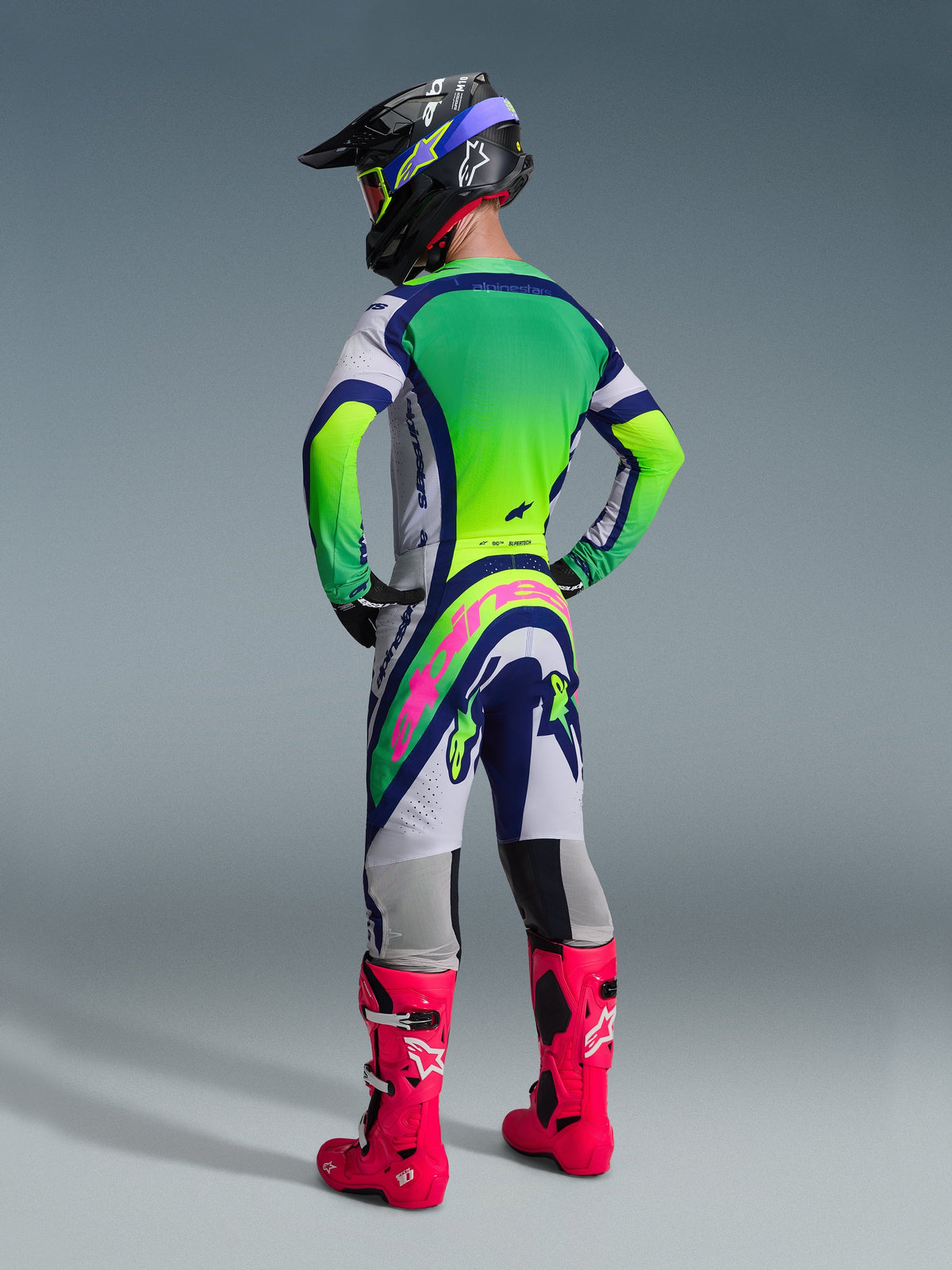 Motocross-Fahrer in der Rückansicht mit der Alpinestars 2026 Supertech Vista Hose, Design in Hellgrau, Blau und Neongelb mit auffälligem pinkfarbenem Logo am Gesäß, dazu passendes Trikot, schwarzer Helm und leuchtend pinkfarbene Stiefel, komplette Offroad-Rennausrüstung.
