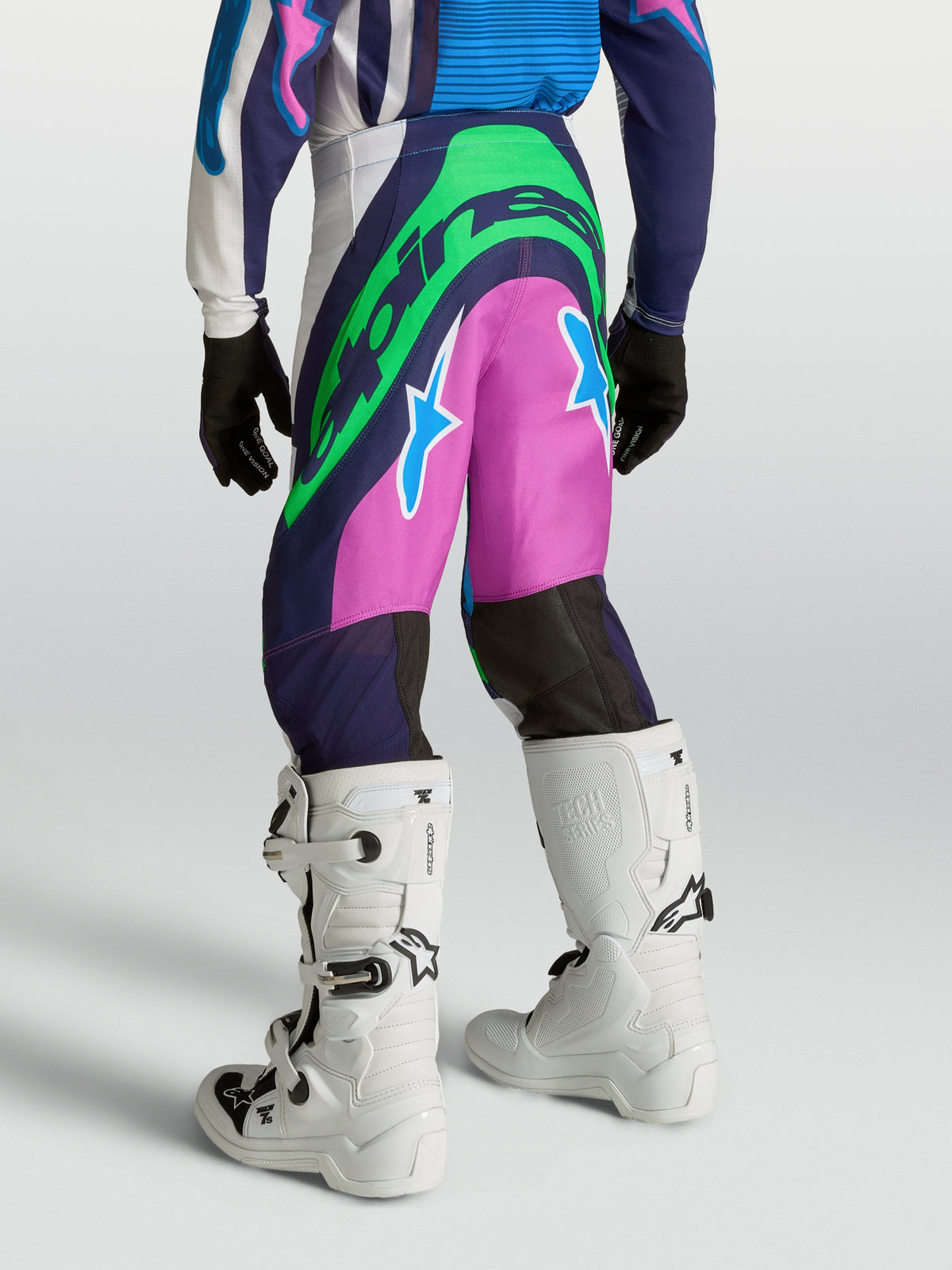 Alpinestars Jugend Supertech Maven Hose, Motocross-Hose in Blau und Pink mit neongrünen Logos, athletische Rennpassform, Rückansicht am Model mit weißen Motocross-Stiefeln für MX-Geländefahrten.
