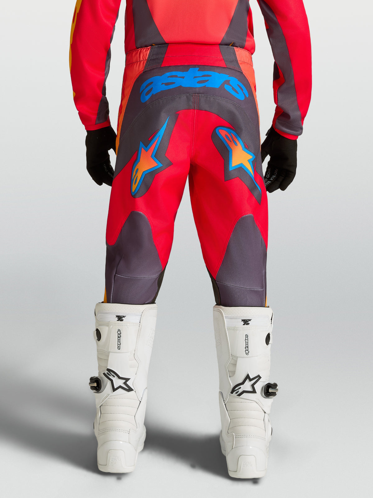 Alpinestars Jugend Fluid Hunt Hose, Motocrosshose in Rot und Grau, Rückansicht mit blauem astars-Branding und Sternengrafiken, strapazierfähiges 600D-Polygewebe, kombiniert mit weißen MX-Stiefeln.