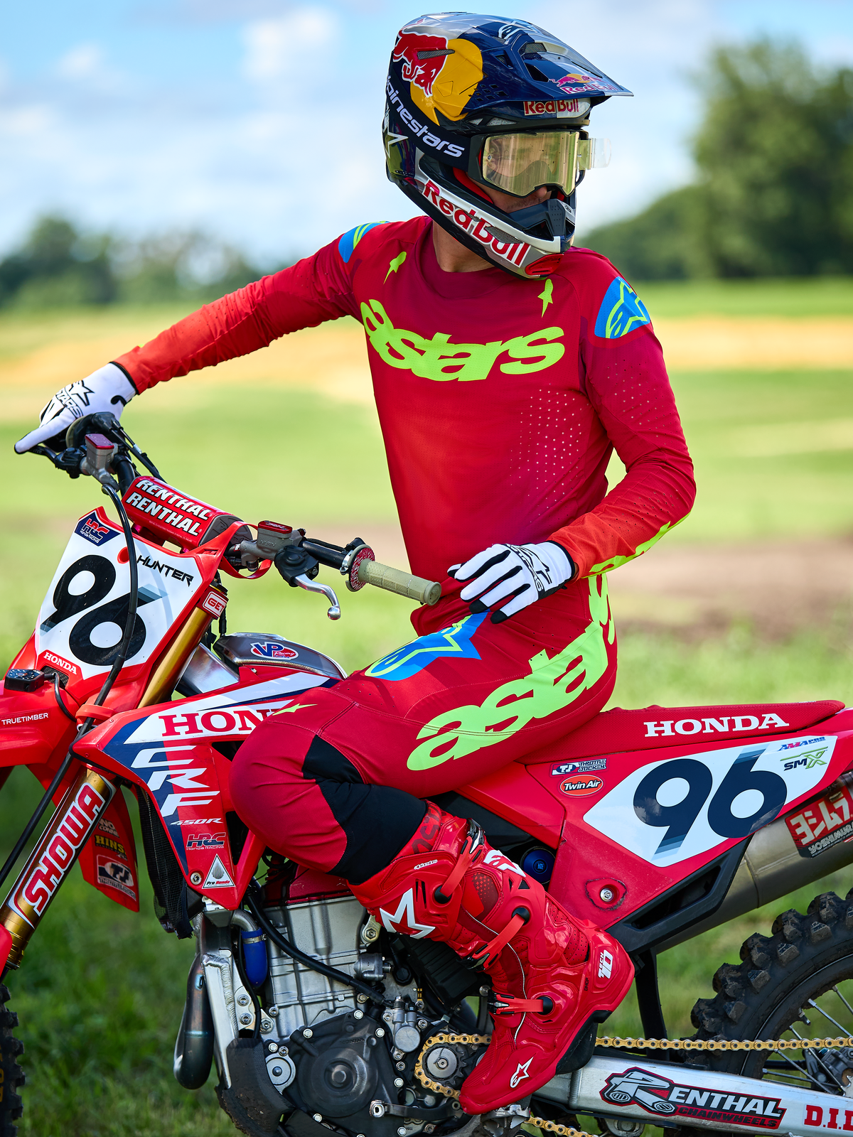 Motocross-Fahrer auf einem roten Honda-Motorrad im Alpinestars 2025 Supertech Maker Gear Set in Rot, bestehend aus einem roten Trikot mit neongrünem „astars“-Branding und blauen Schulterakzenten, passender roter Hose, roten Stiefeln, weißen Handschuhen und einem Red Bull-Helm, professionelle Rennausrüstung für Offroad-Performance
