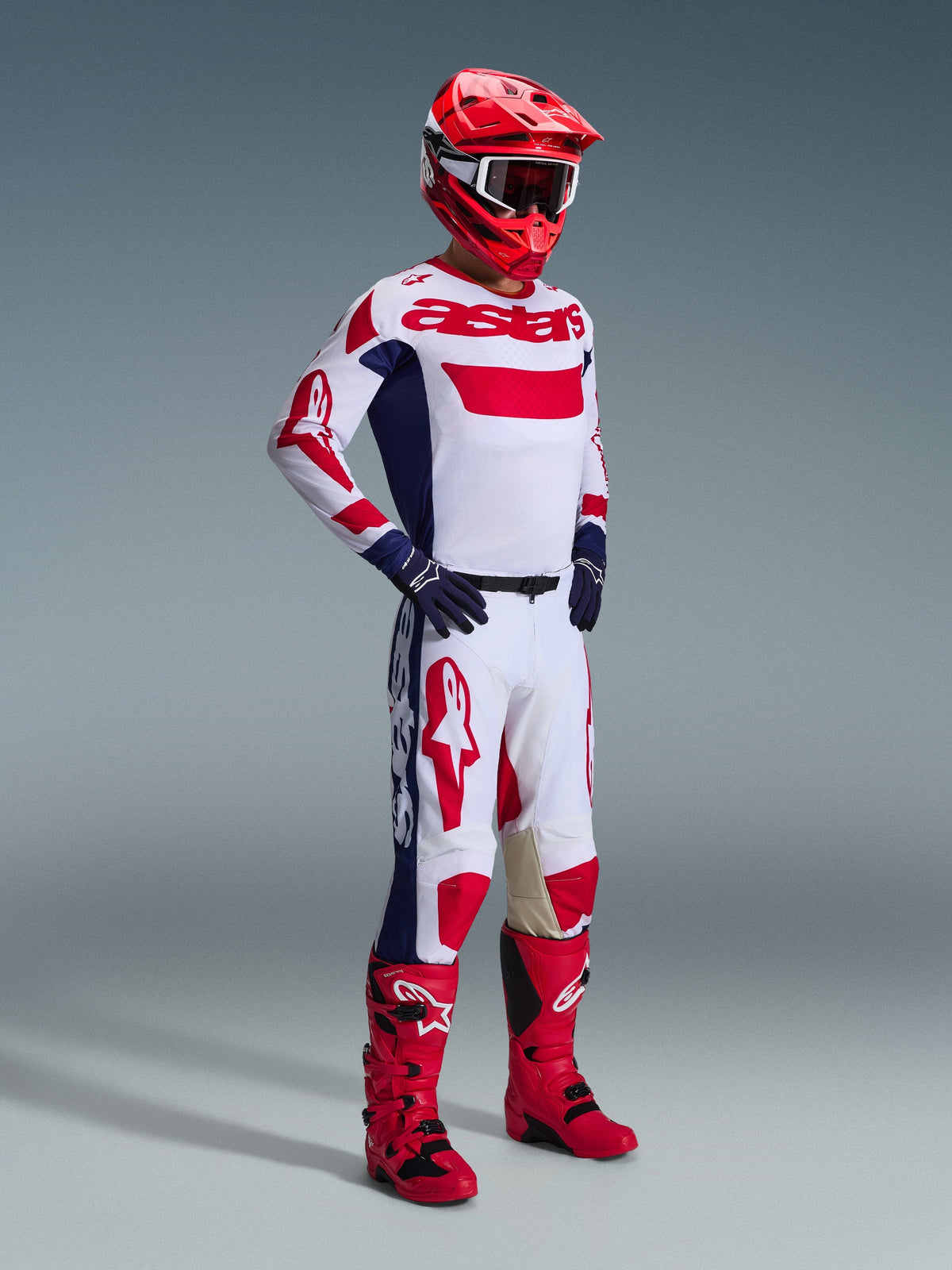 Motocross-Fahrer im Alpinestars 2026 Racer Riway Gear Set, weißes Trikot mit roten und blauen Akzenten und 'astars'-Branding, passende weiße Hose mit großen roten Logos und dunkelblauen Seiteneinsätzen, roter Helm, rot-schwarze Stiefel und dunkelblaue Handschuhe, professionelle Rennausrüstung für Offroad-Performance.
