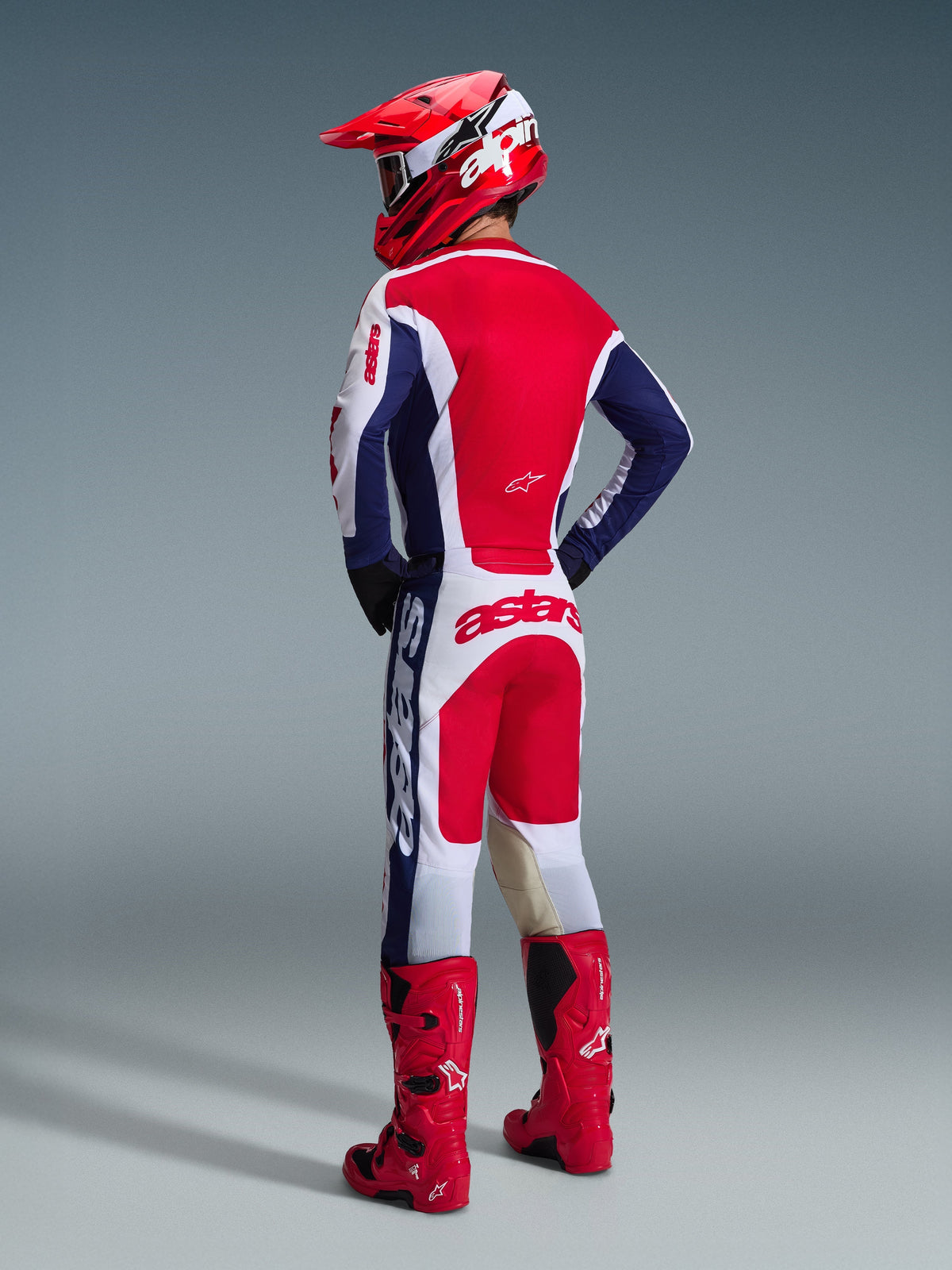 Motocross-Fahrer von hinten gesehen, trägt das Alpinestars 2026 Racer Riway Ausrüstungsset in Weiß-Blau-Rot, bestehend aus einem langärmeligen Trikot mit Astars-Branding und passender Hose, kombiniert mit einem roten Helm und roten Offroad-Stiefeln.